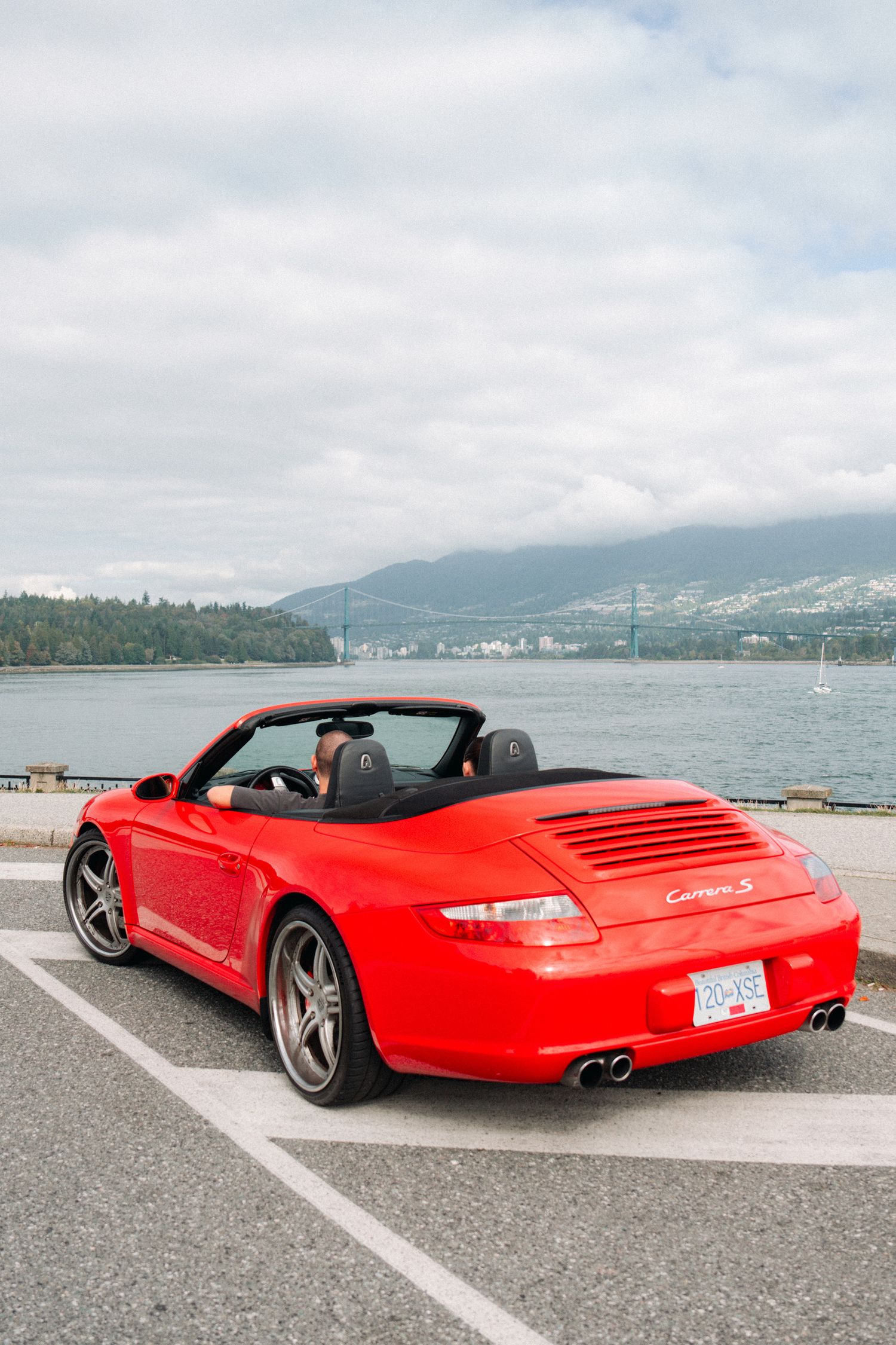 Red Porsche 911 convertible parked by a waterfront with mountains in the background.