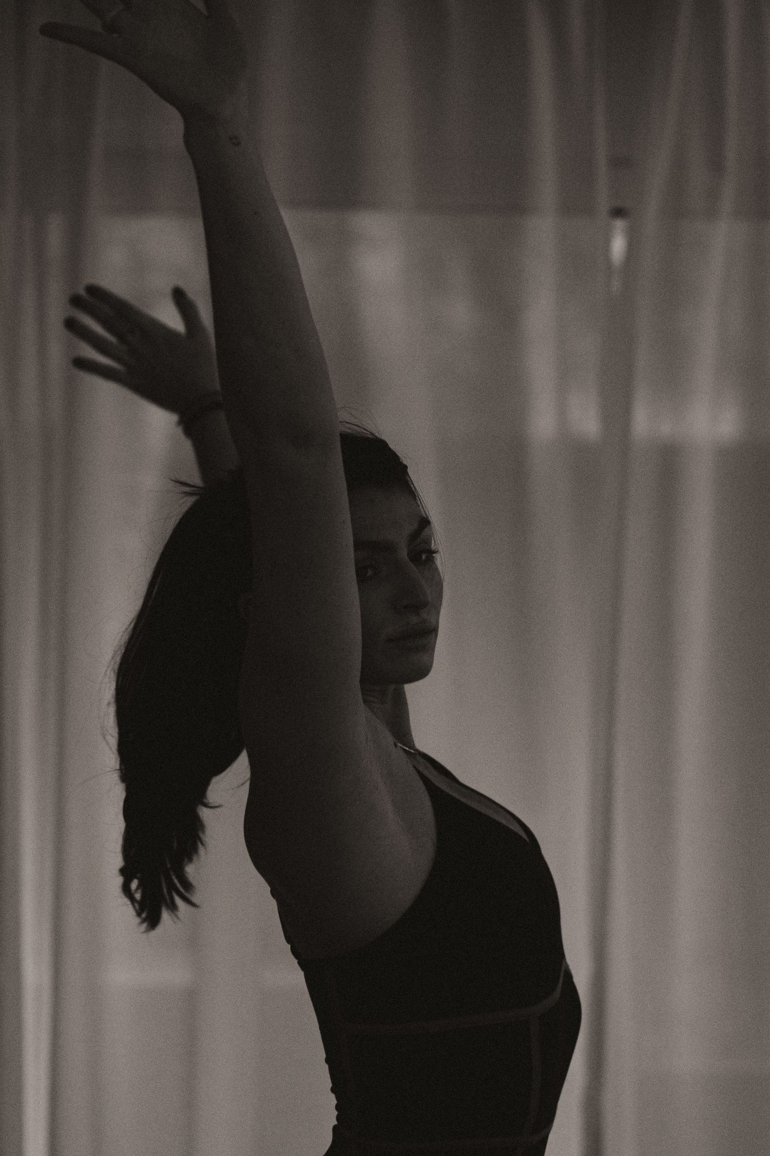 A silhouetted dancer strikes an elegant pose with raised arm against soft curtains in moody black and white lighting.