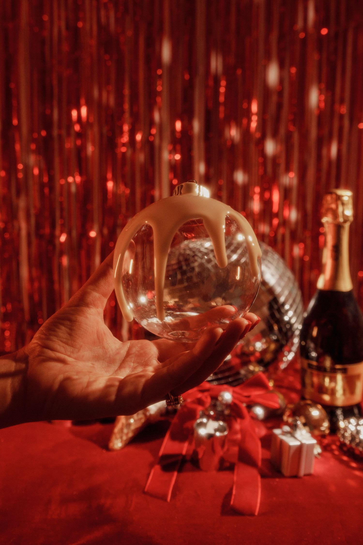 Hand holding clear glass ornament against red sparkly background with champagne and gifts.
