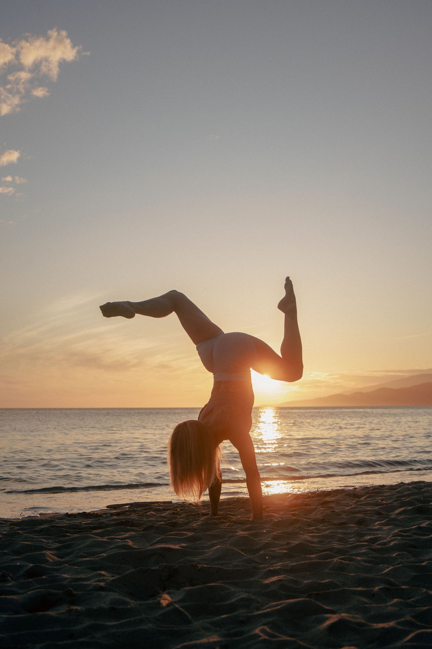 Gymnast performs handstand on beach with setting sun creating dramatic silhouette.