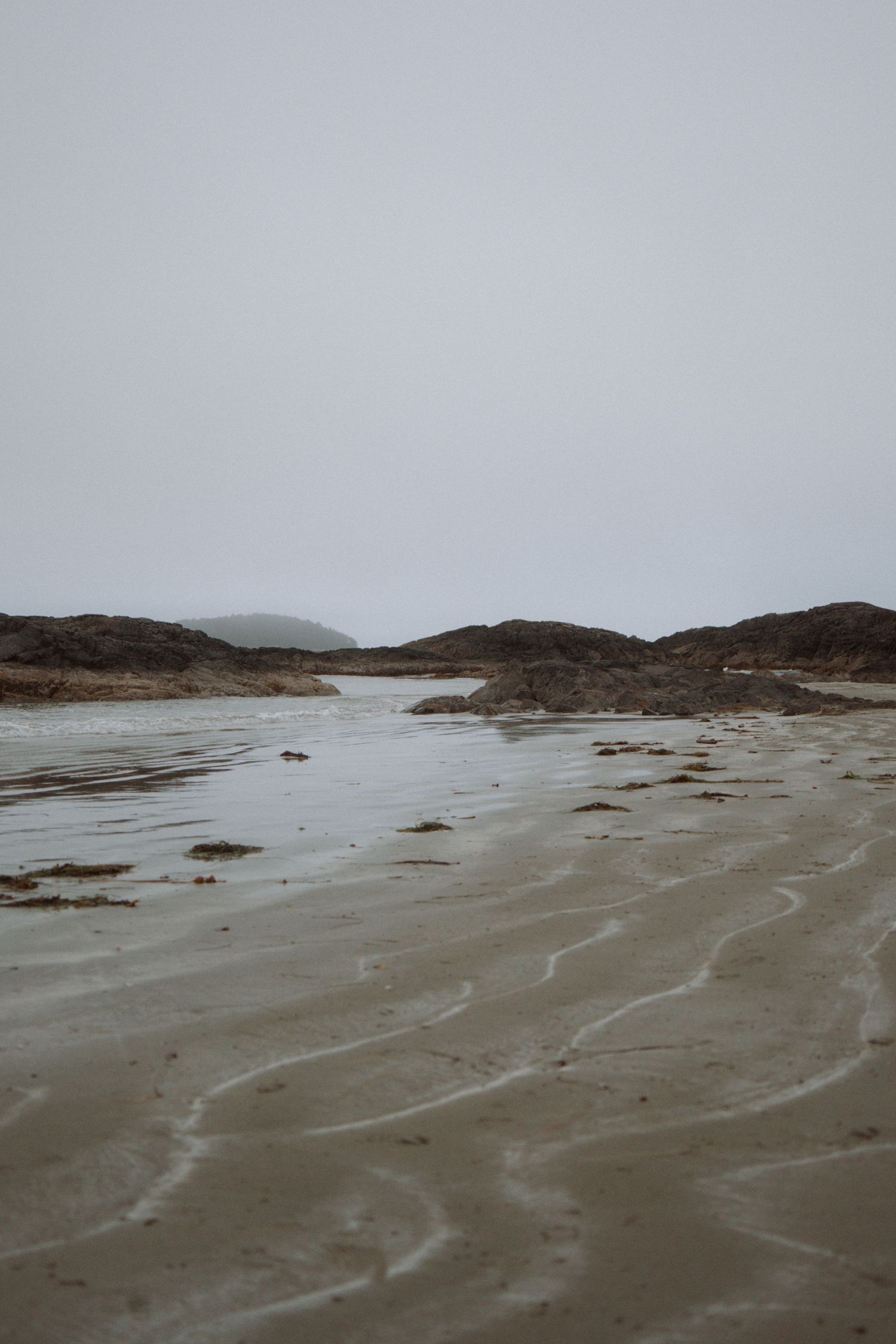 Moody coastal landscape with rippled wet sand and rocky outcrops under overcast sky on a misty beach.