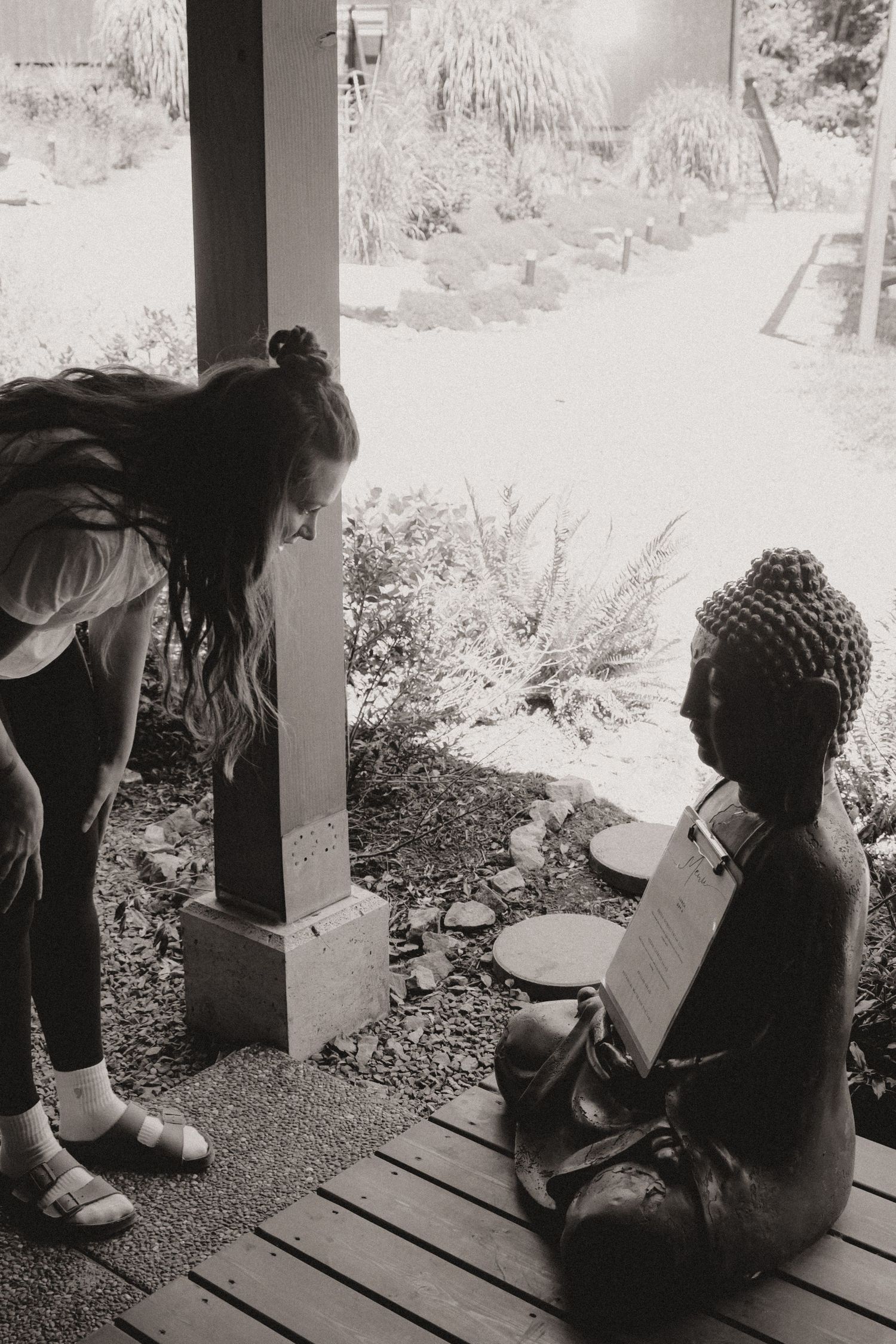 Black and white photo of someone bending down near a decorative Buddha statue on a porch.
