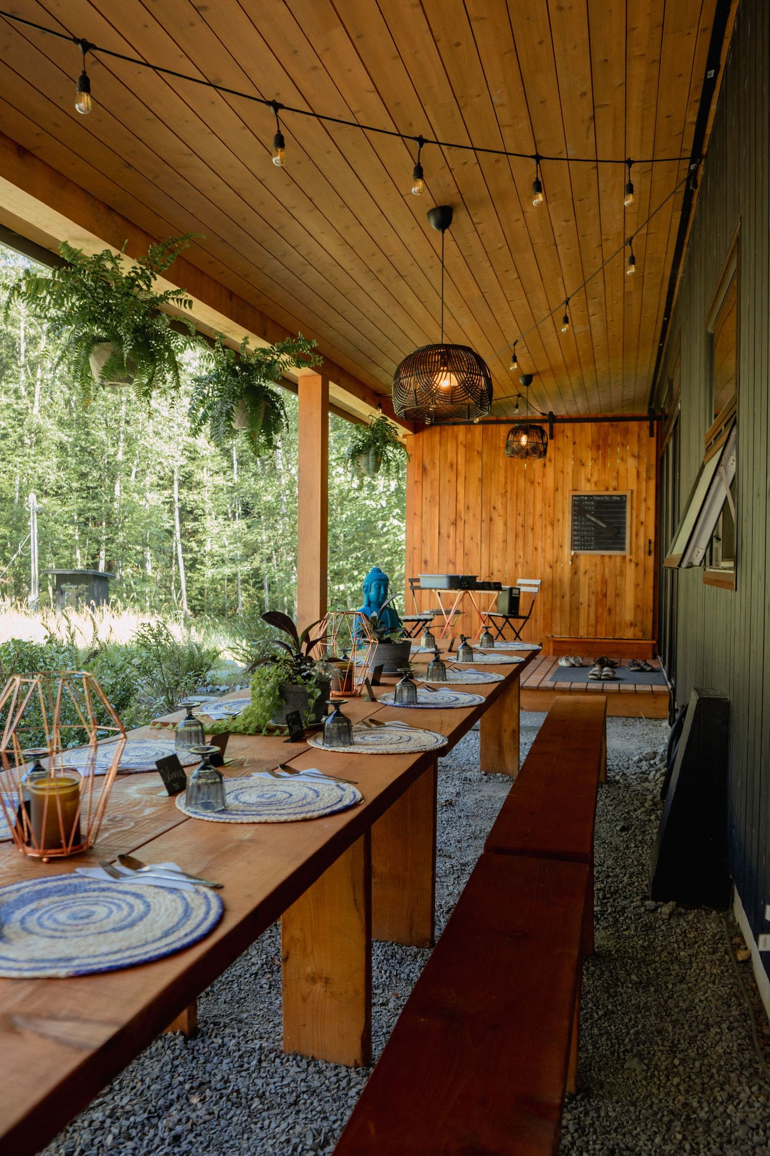 Outdoor dining area with wooden tables and string lights under a covered porch in the forest.