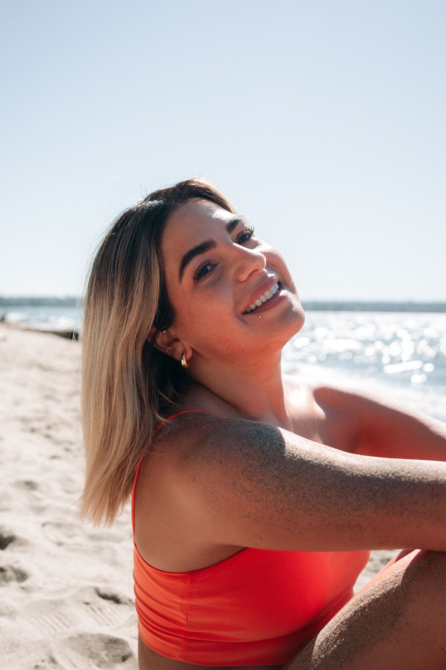 Woman in orange swimsuit smiling on sunny beach with ocean in background.