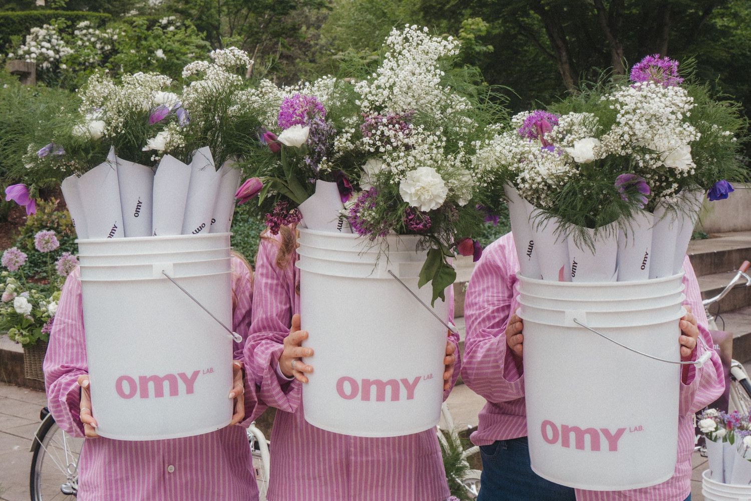 Three white metal buckets with pink 'omy' logos filled with delicate white and pink floral arrangements.