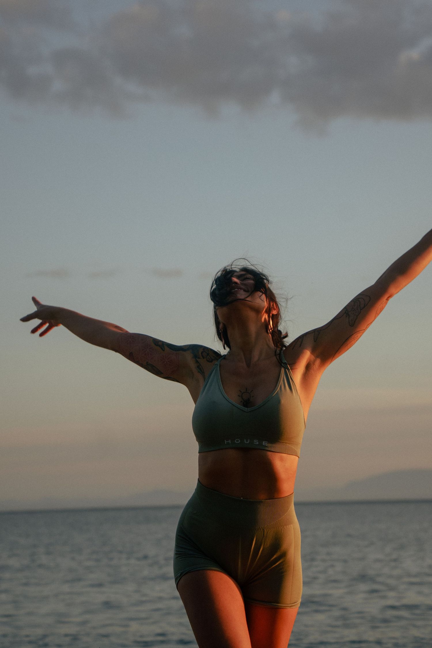 Silhouetted figure in athletic wear stretches joyfully with arms outstretched against sunset ocean backdrop.