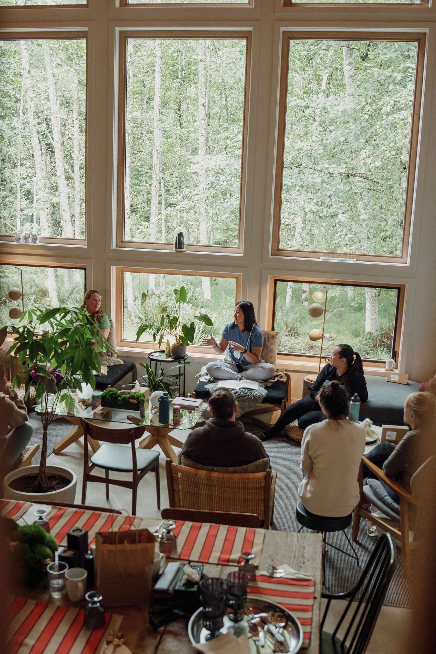 A cozy living room gathering with large windows overlooking a forest, featuring houseplants and a striped rug.