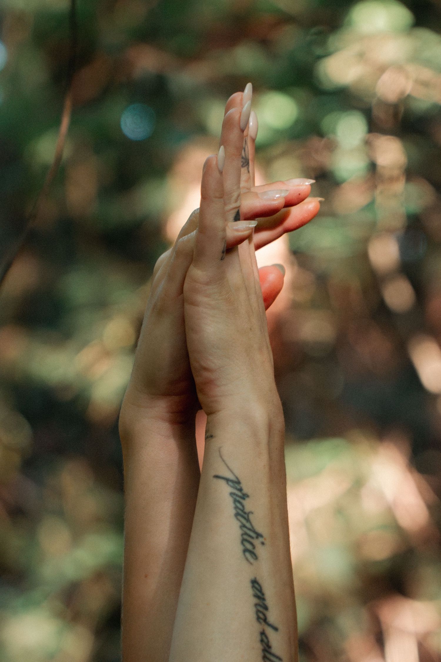 Close-up of hands pressed together against blurred forest background, showing script tattoo.