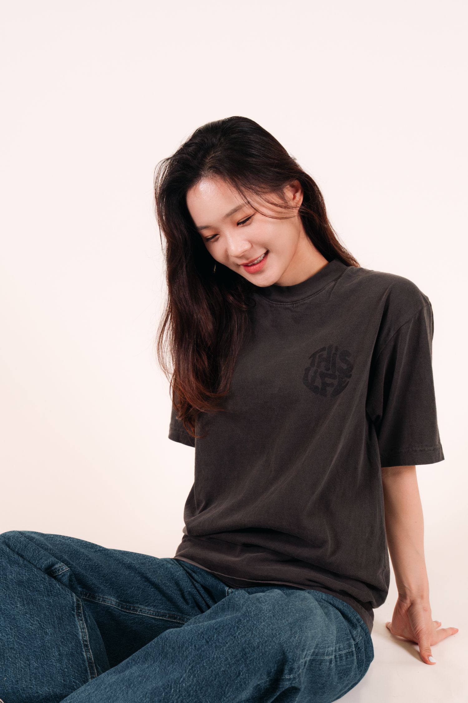 Model wearing dark t-shirt with logo smiles while sitting casually against white background.