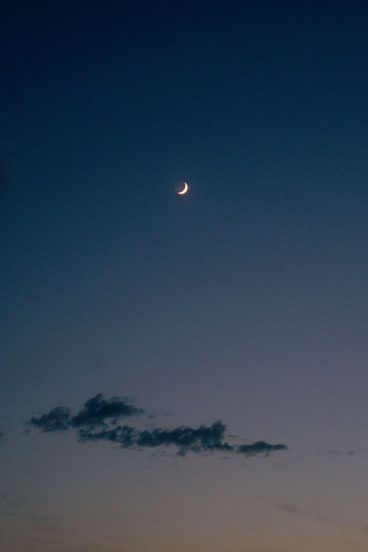Crescent moon in dark blue evening sky with wispy clouds below.