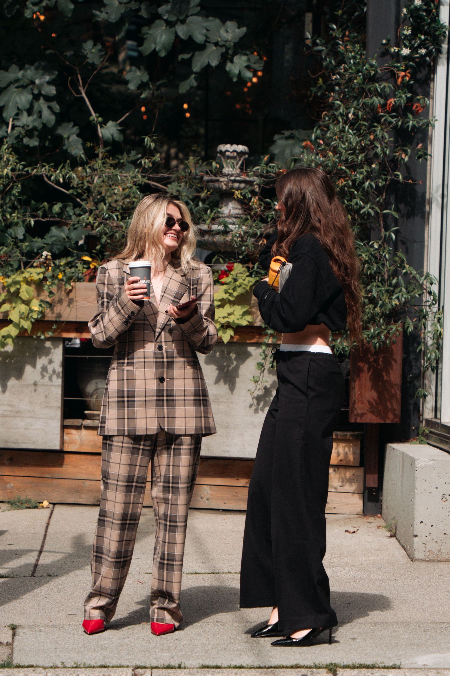 Two friends in trendy outfits enjoying coffee outside on a sunny day.