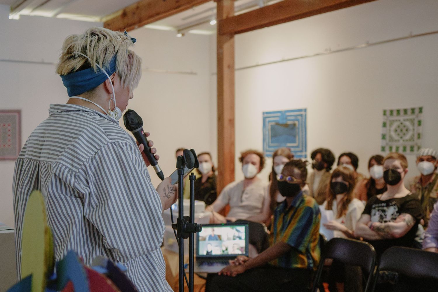A performer addresses a seated audience in a bright room with wooden beams.