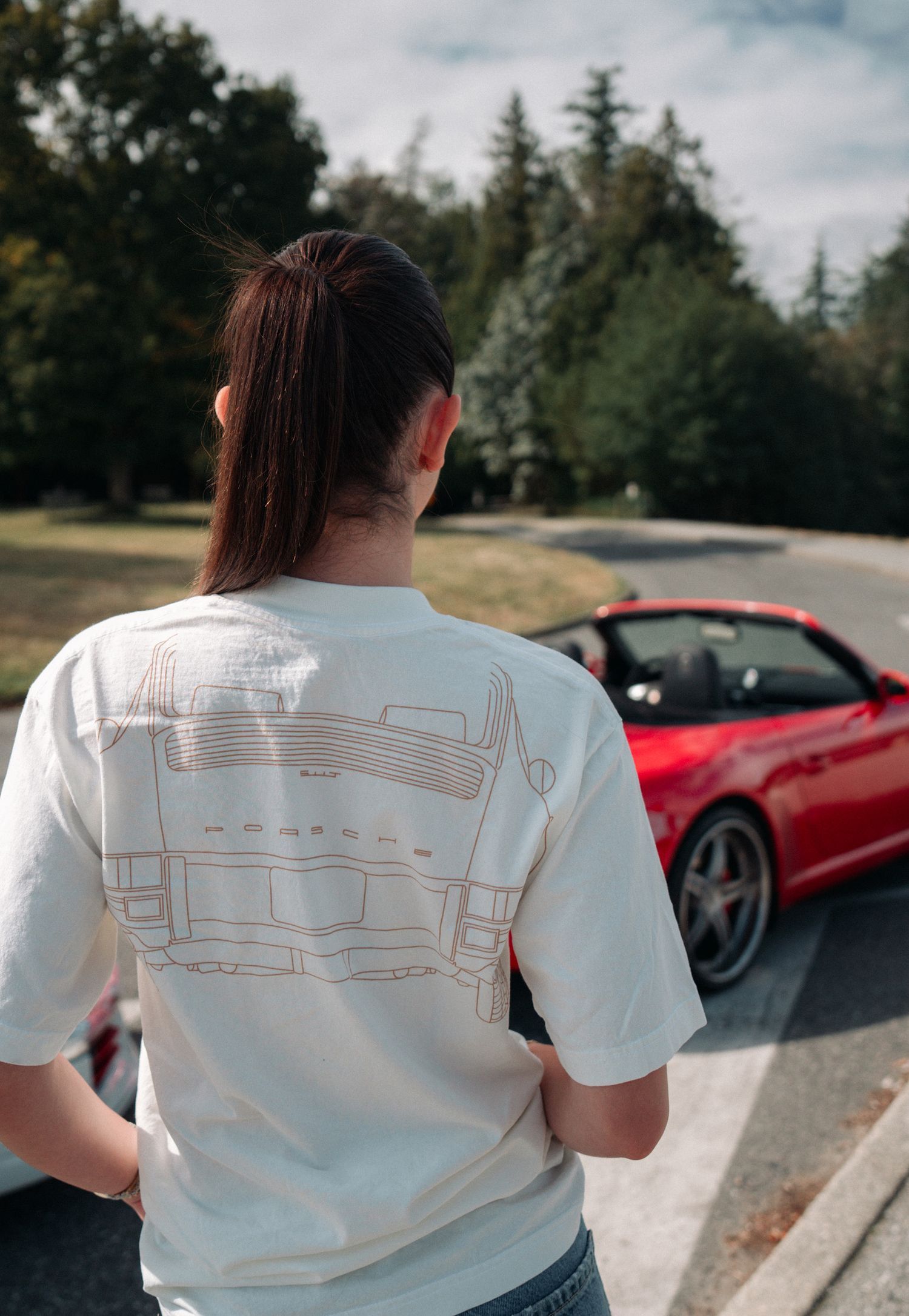 Back view of someone wearing a white t-shirt with blueprint design standing near a red sports car.
