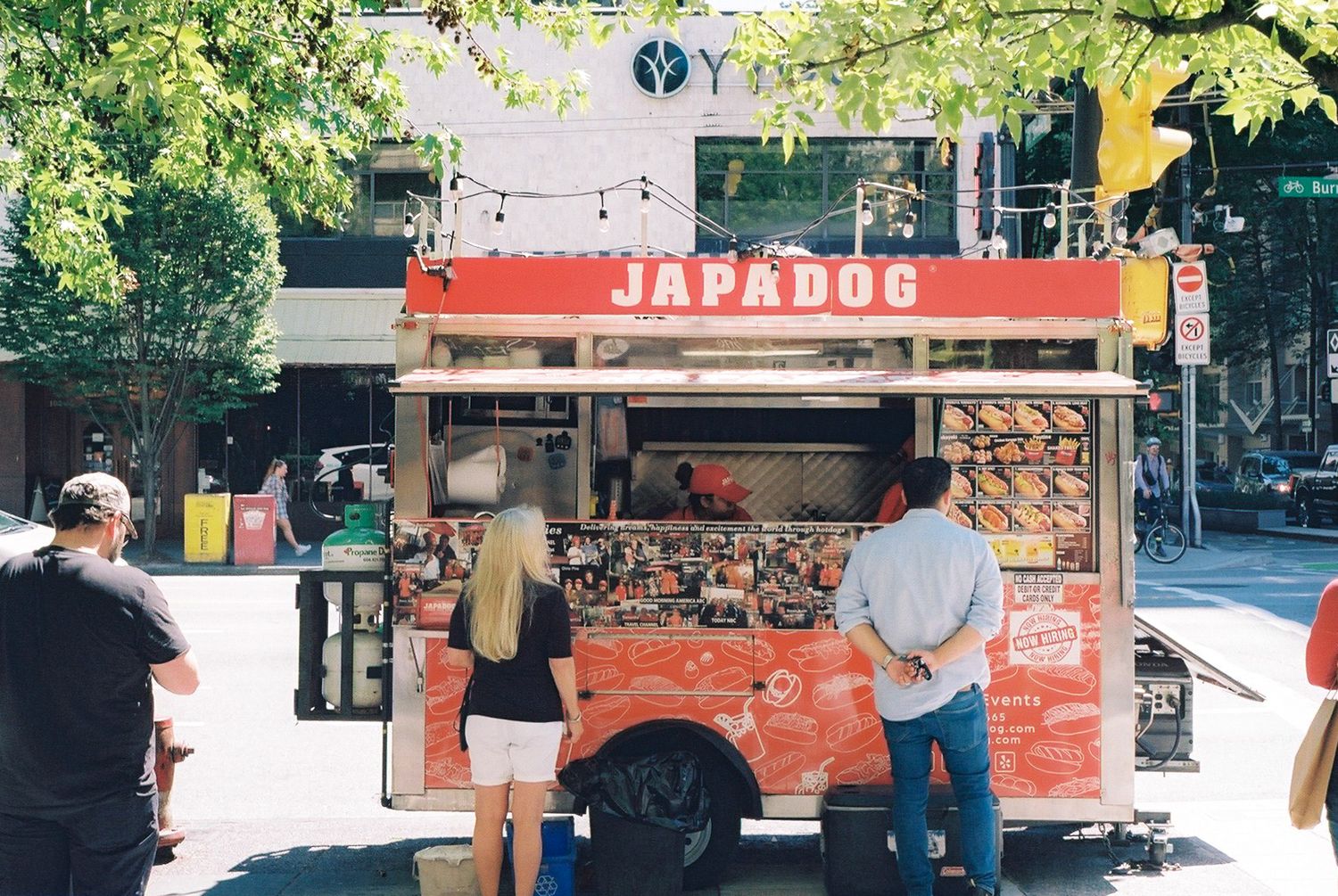 Red Japadog food cart with customers waiting in line on a city street.