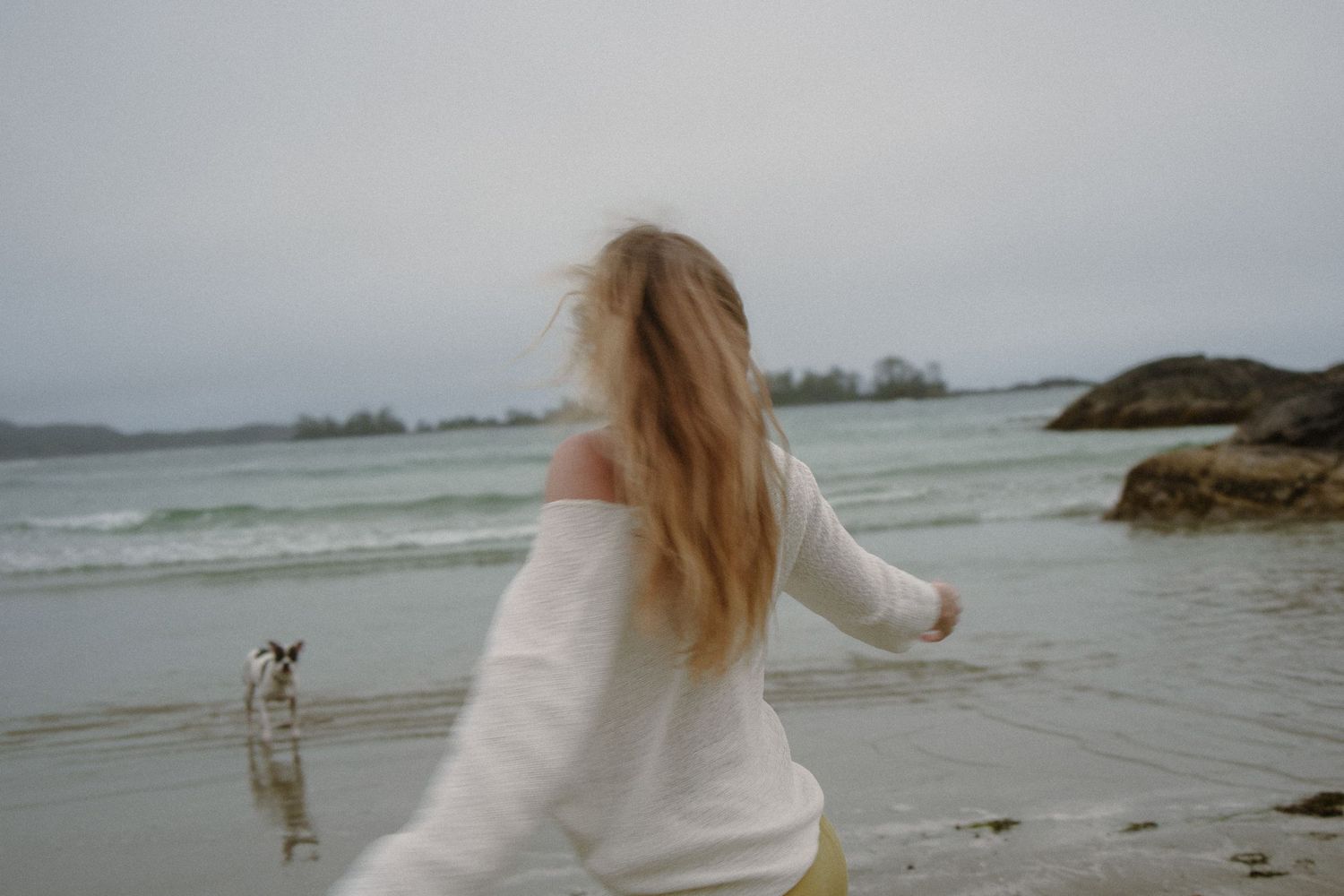 A person in a white sweater spins joyfully on a cloudy beach while a small dog watches nearby.