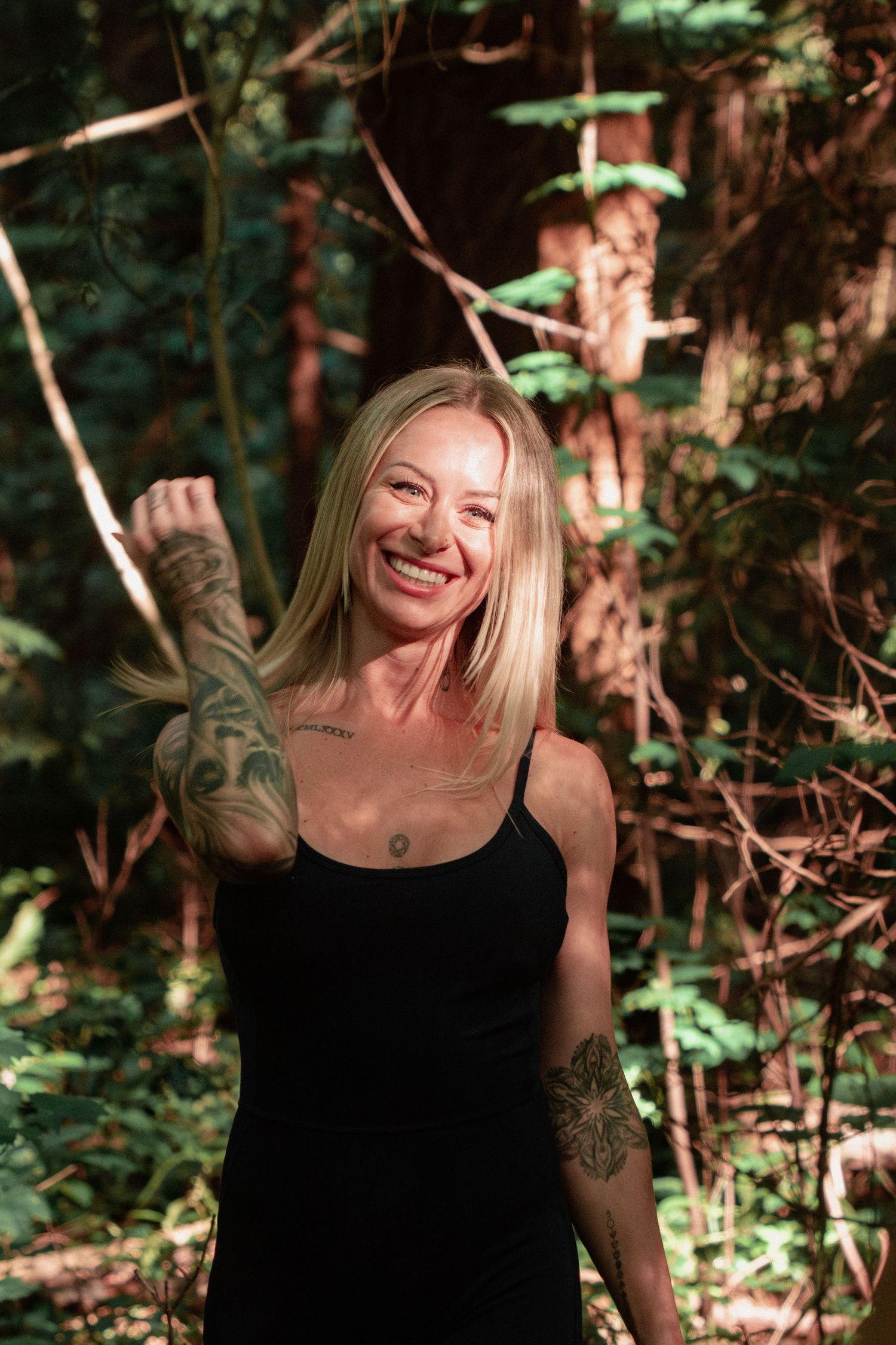 Woman in black tank top smiling joyfully in a sunlit forest setting.