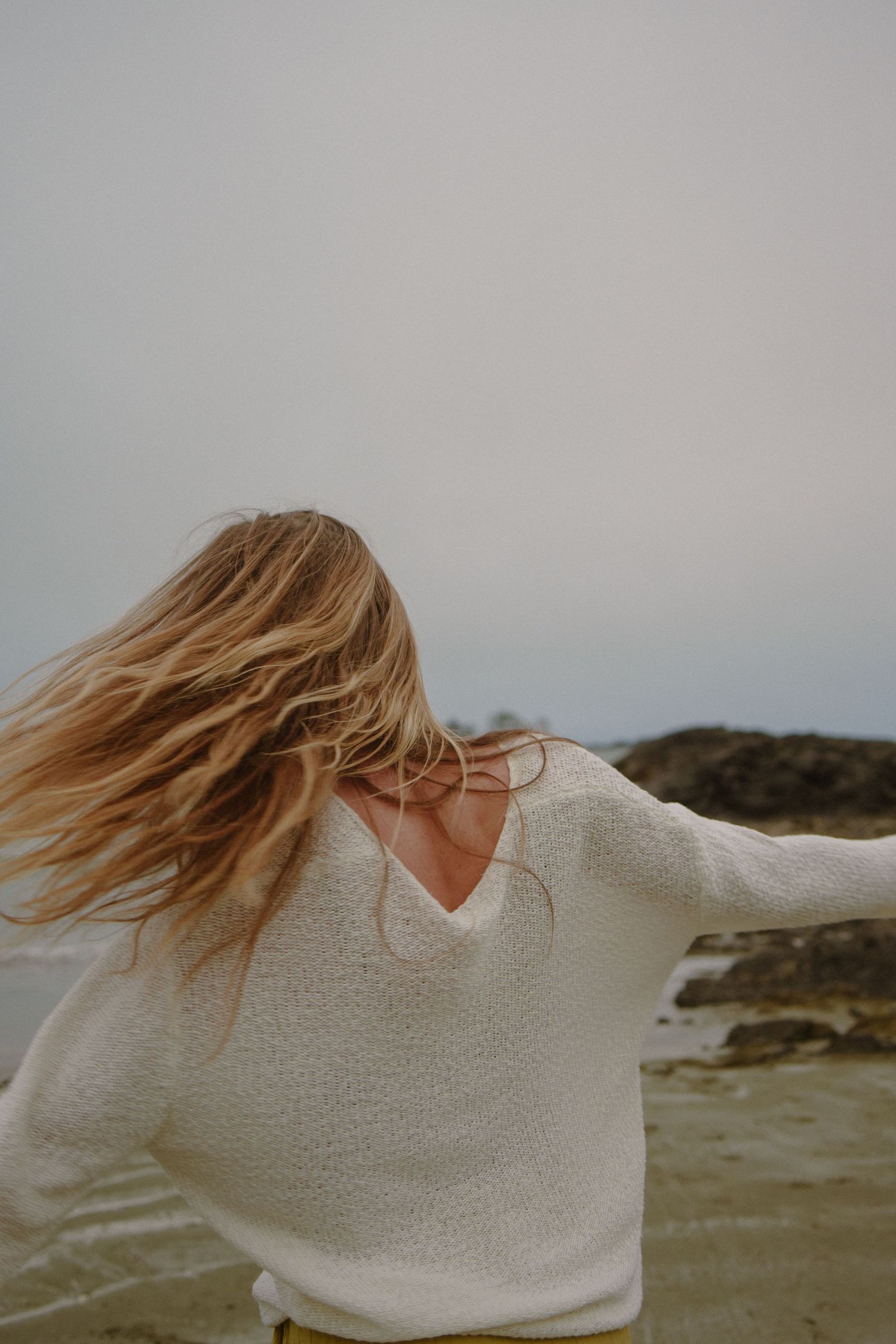 Person in white sweater spinning with windswept hair on beach.