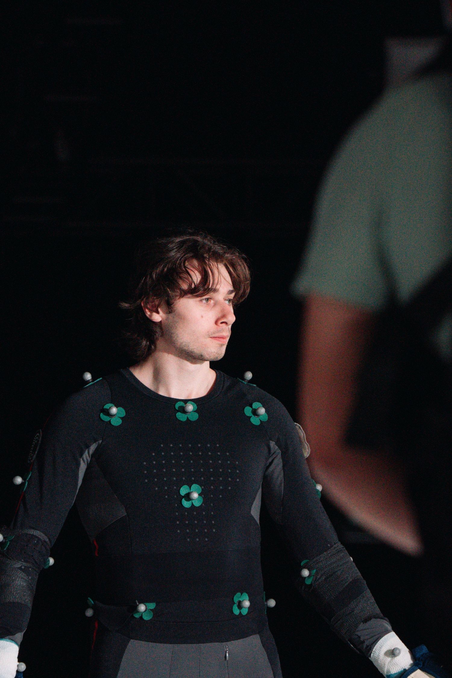 Actor wearing a black motion capture suit with green markers in dramatic studio lighting.