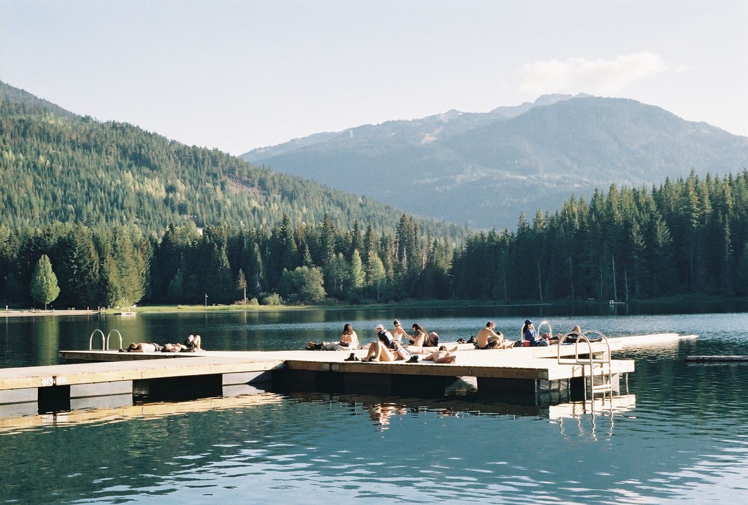 People relaxing on a wooden dock by a serene mountain lake surrounded by evergreen forests on a sunny day.