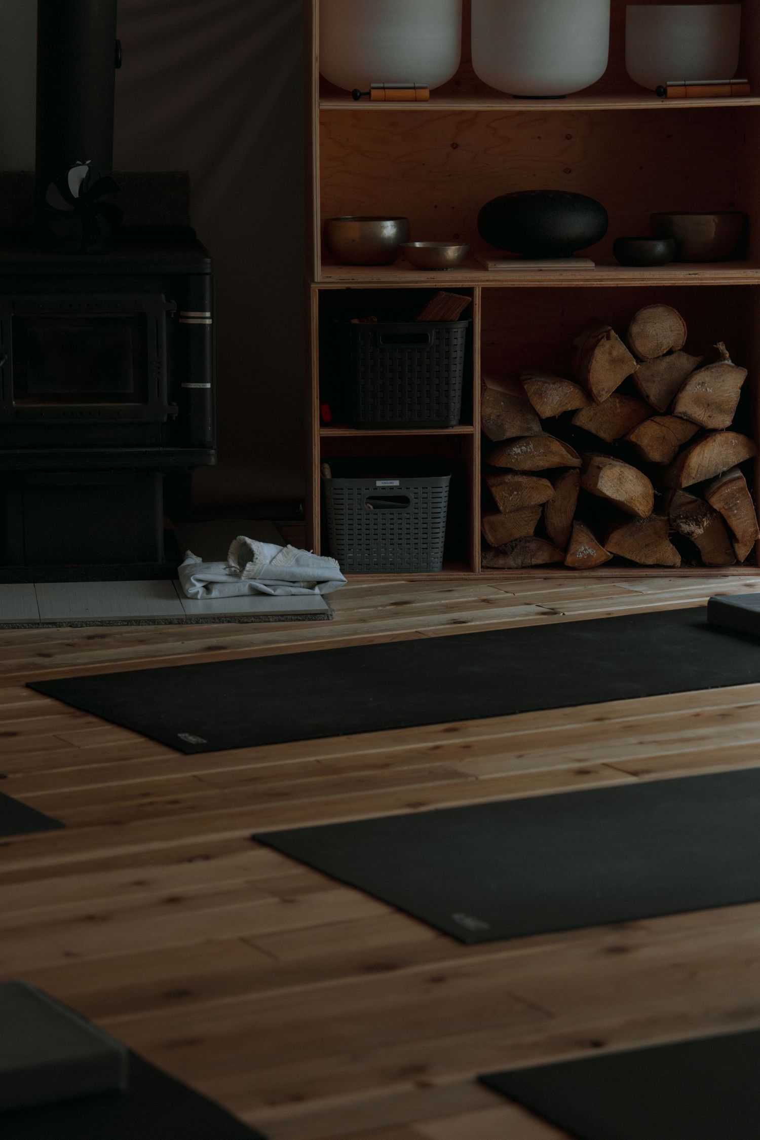 Yoga studio with black mats on wooden floor next to wood-burning stove and firewood storage.