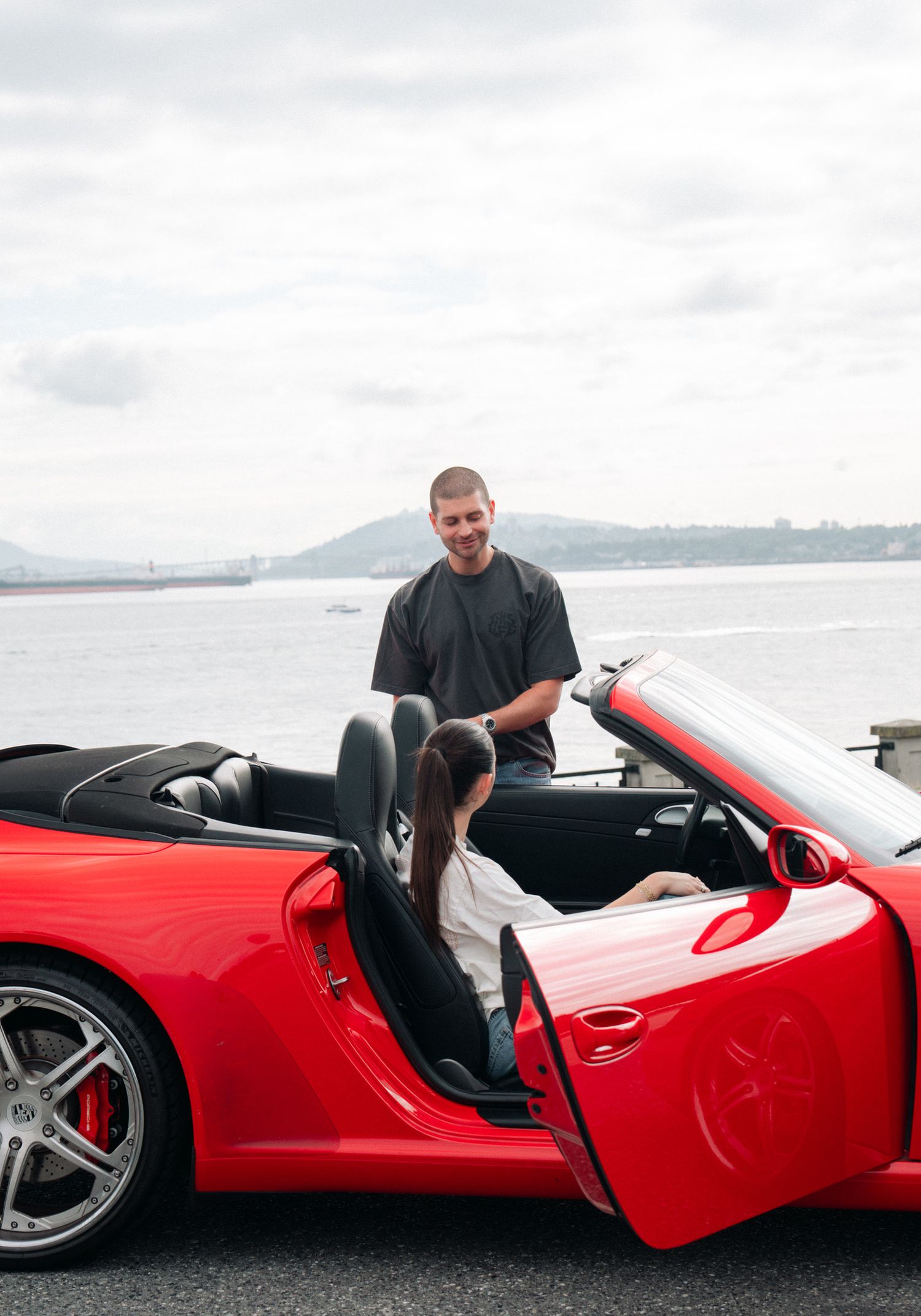 A red convertible sports car parked near a waterfront with mountains in the background.