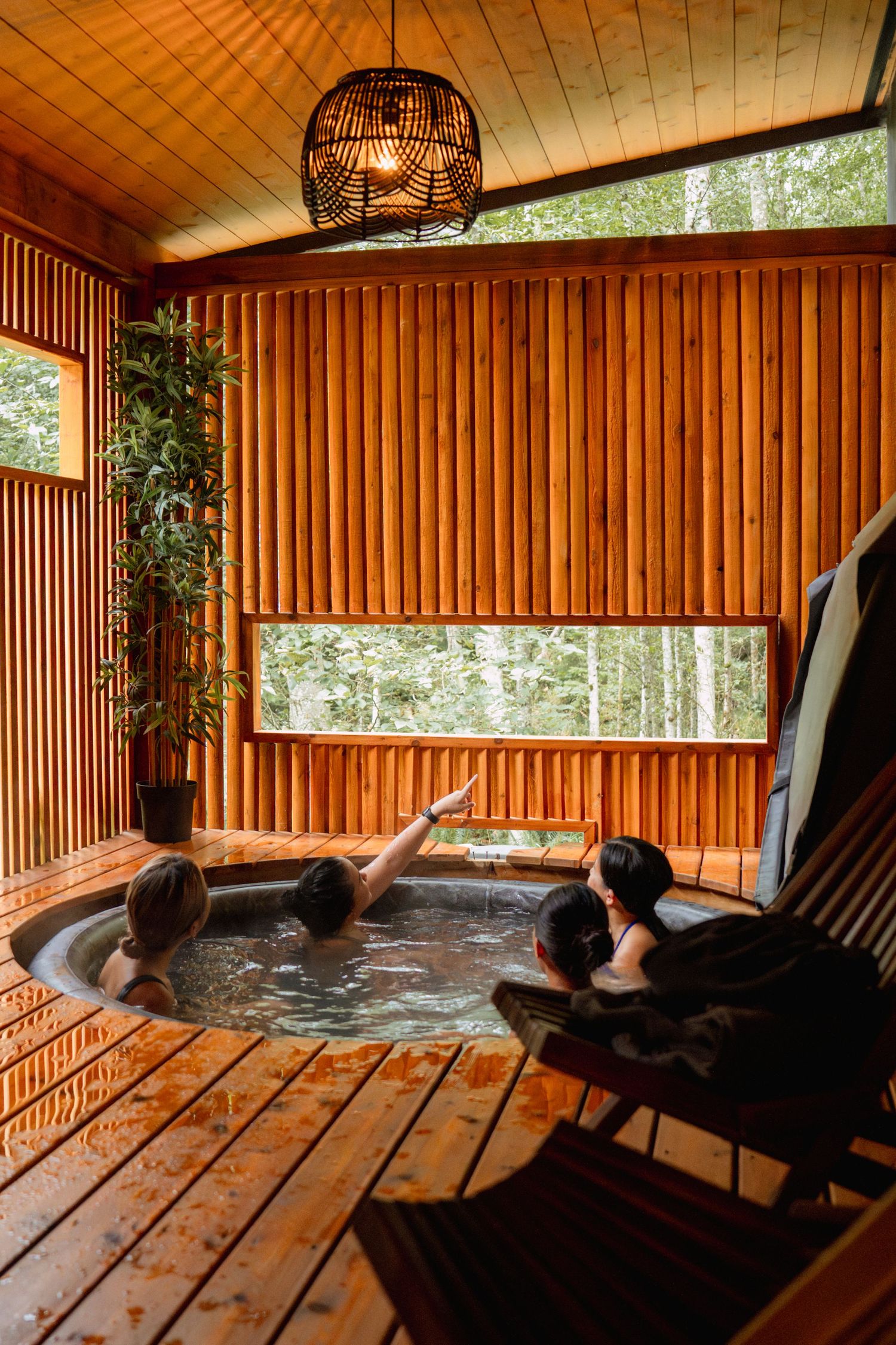 Hot tub surrounded by wooden walls and wicker pendant light with forest view through window.