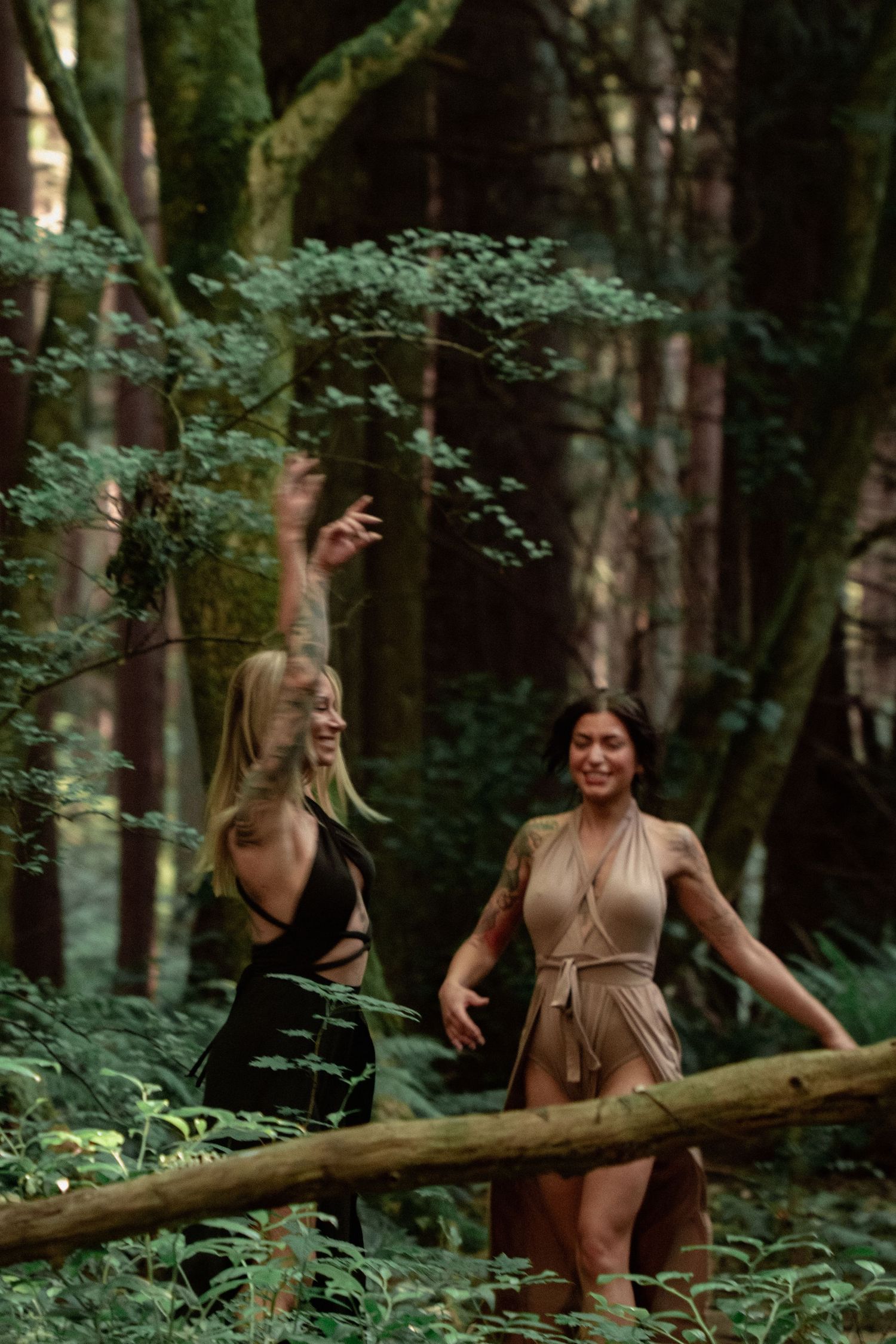 Two people in stylish dresses share a playful moment while balancing on a log in a lush forest.