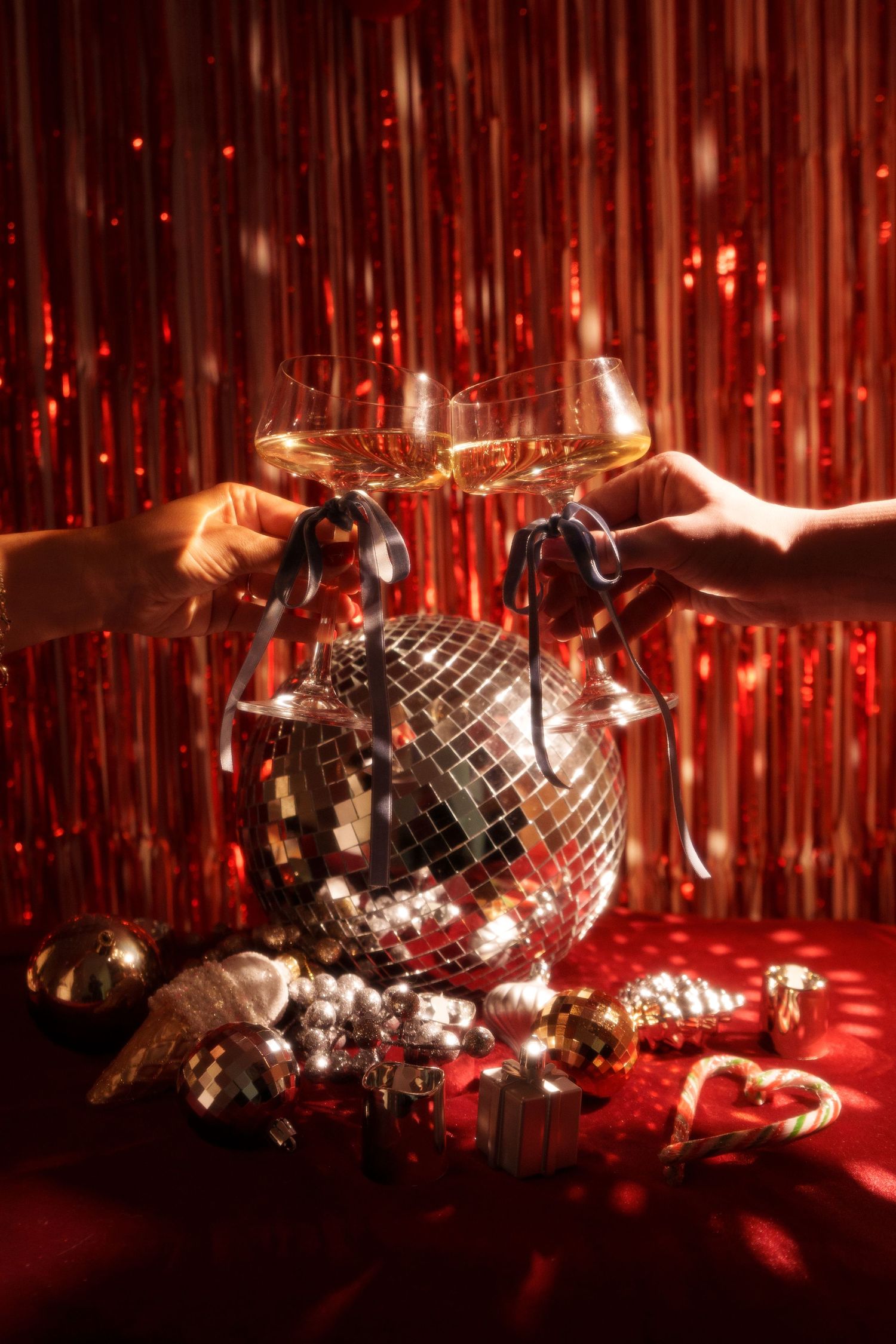 Two hands toasting champagne glasses over disco ball with festive holiday decorations.