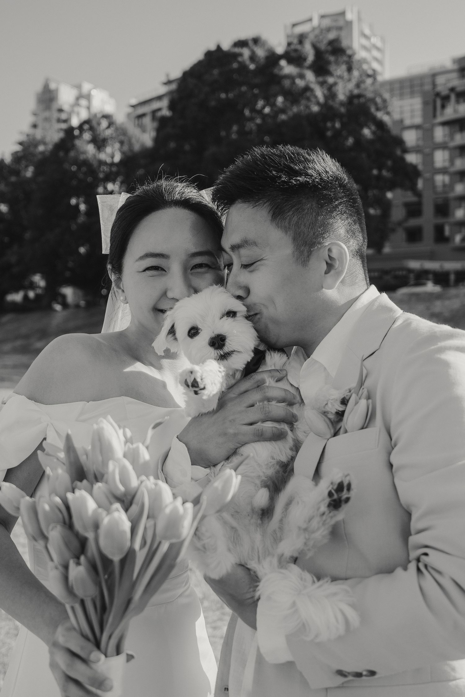 Wedding couple holding small white dog with bouquet of tulips in urban park setting.
