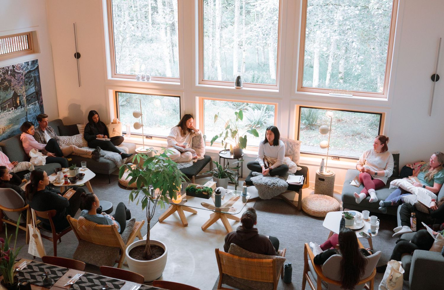 A bright room with floor-to-ceiling windows where people gather on comfortable seating surrounded by plants.
