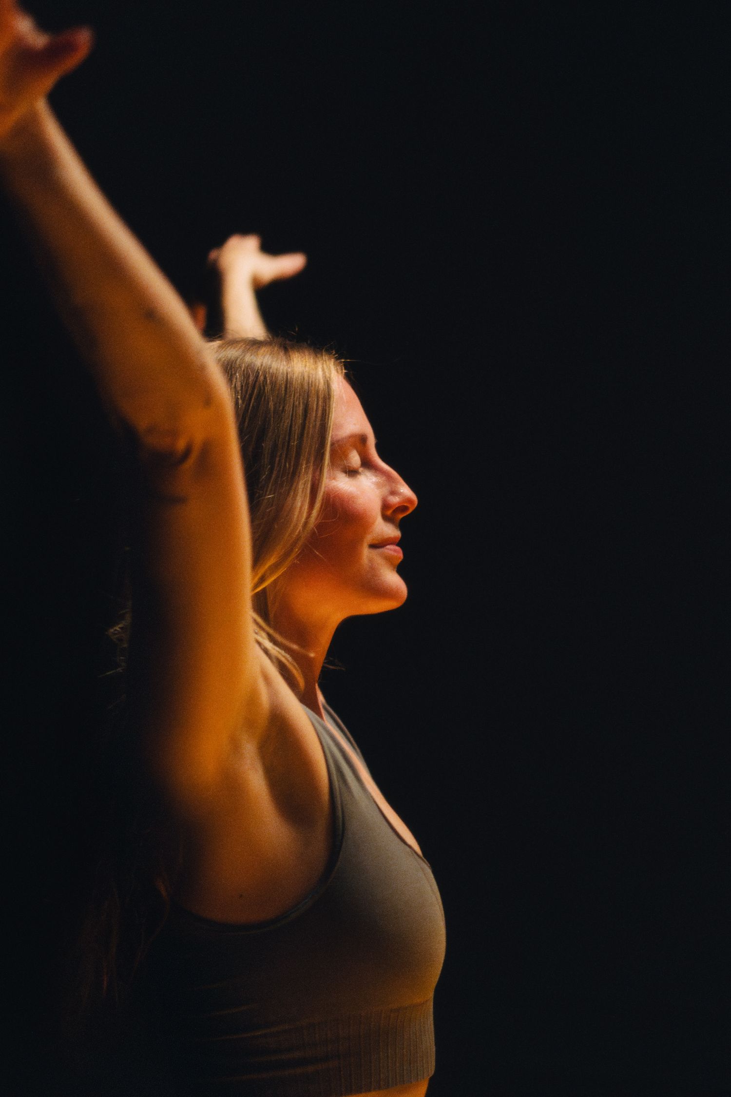 Expressive dancer raises arms in celebration against dark backdrop with warm lighting.