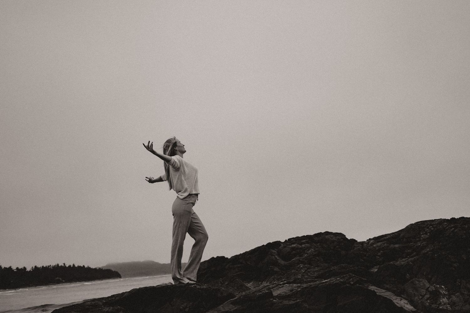 Figure leaping with outstretched arms on a rocky coastal outcrop against a moody sky.