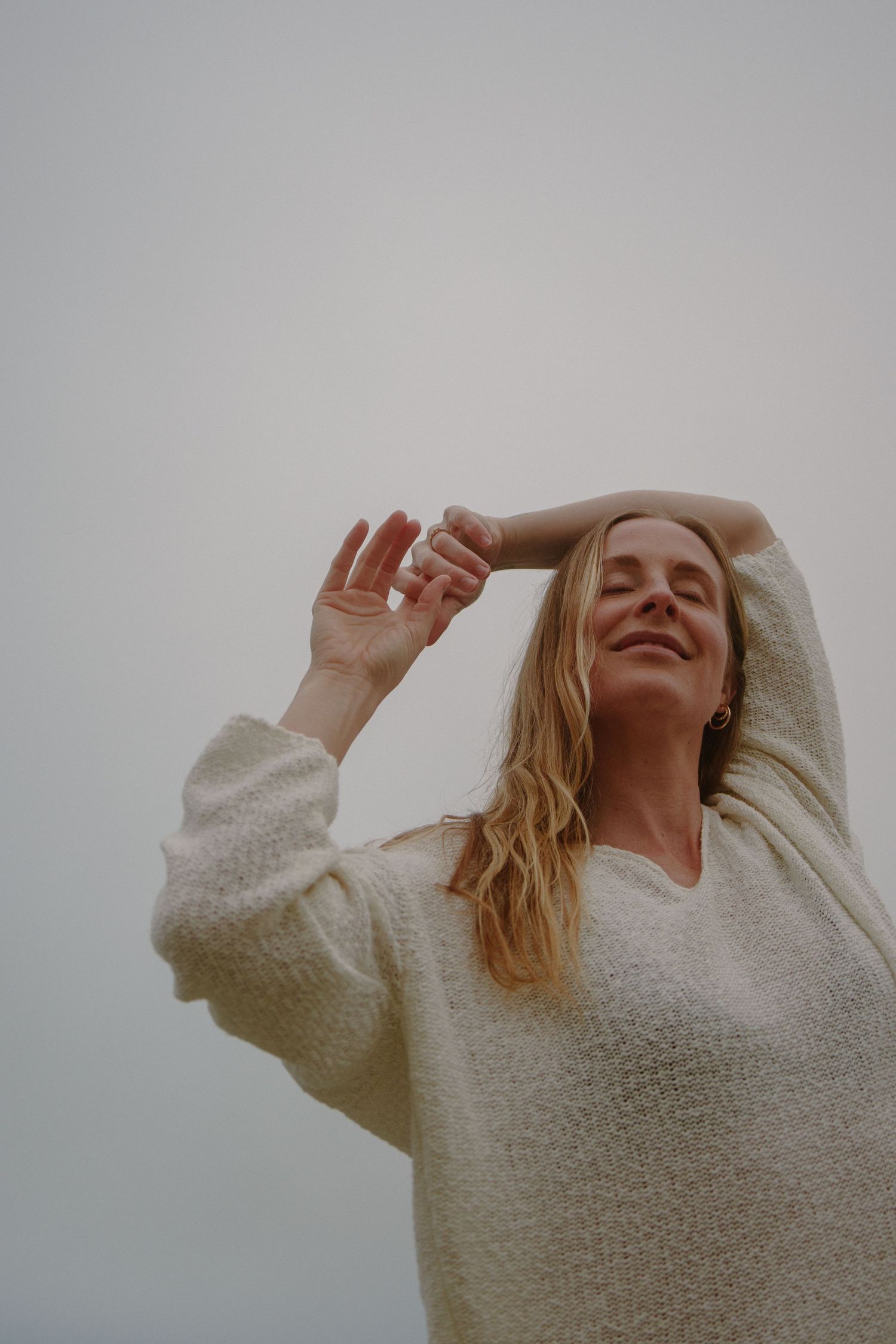 Woman in cozy white sweater poses with raised arms against cloudy sky.