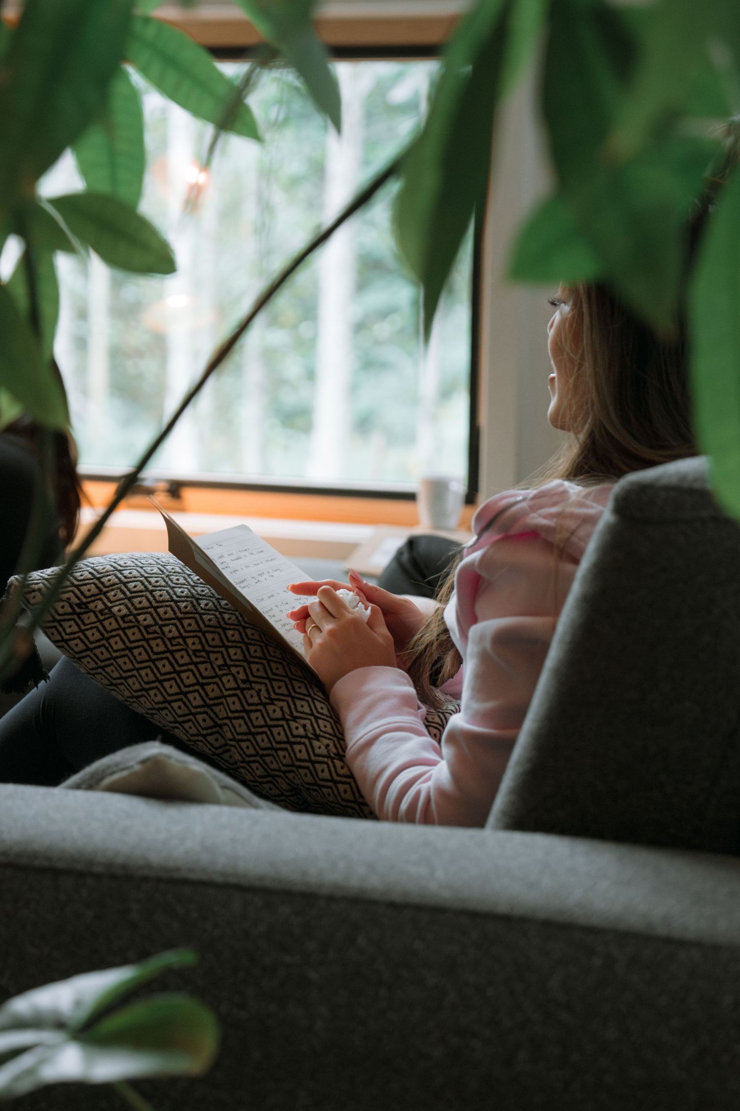 A person lounging on a couch near large windows with leafy plants in the foreground.