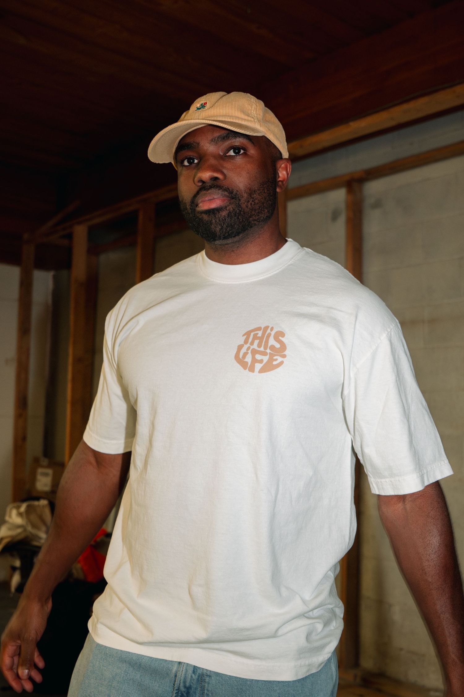 Person wearing tan cap and white t-shirt poses in front of wooden beams.