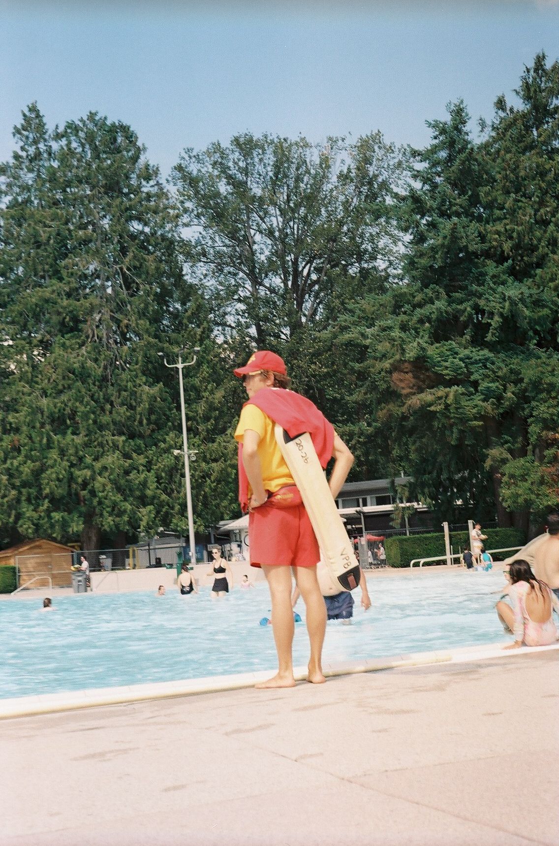 Lifeguard standing at edge of crowded outdoor swimming pool on sunny day.