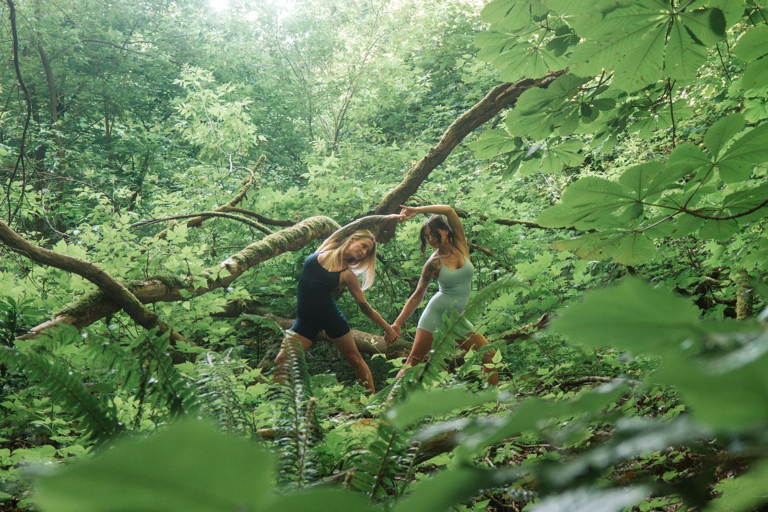 Two dancers performing artistic movements on a fallen tree branch in a lush green forest setting.