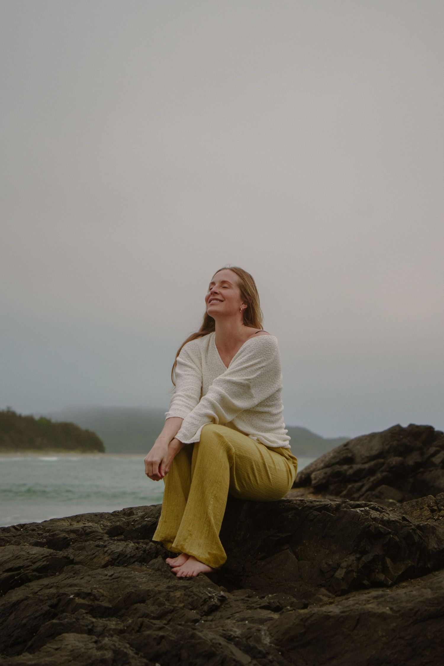 Person in cream sweater and yellow pants sitting on coastal rocks overlooking the ocean.