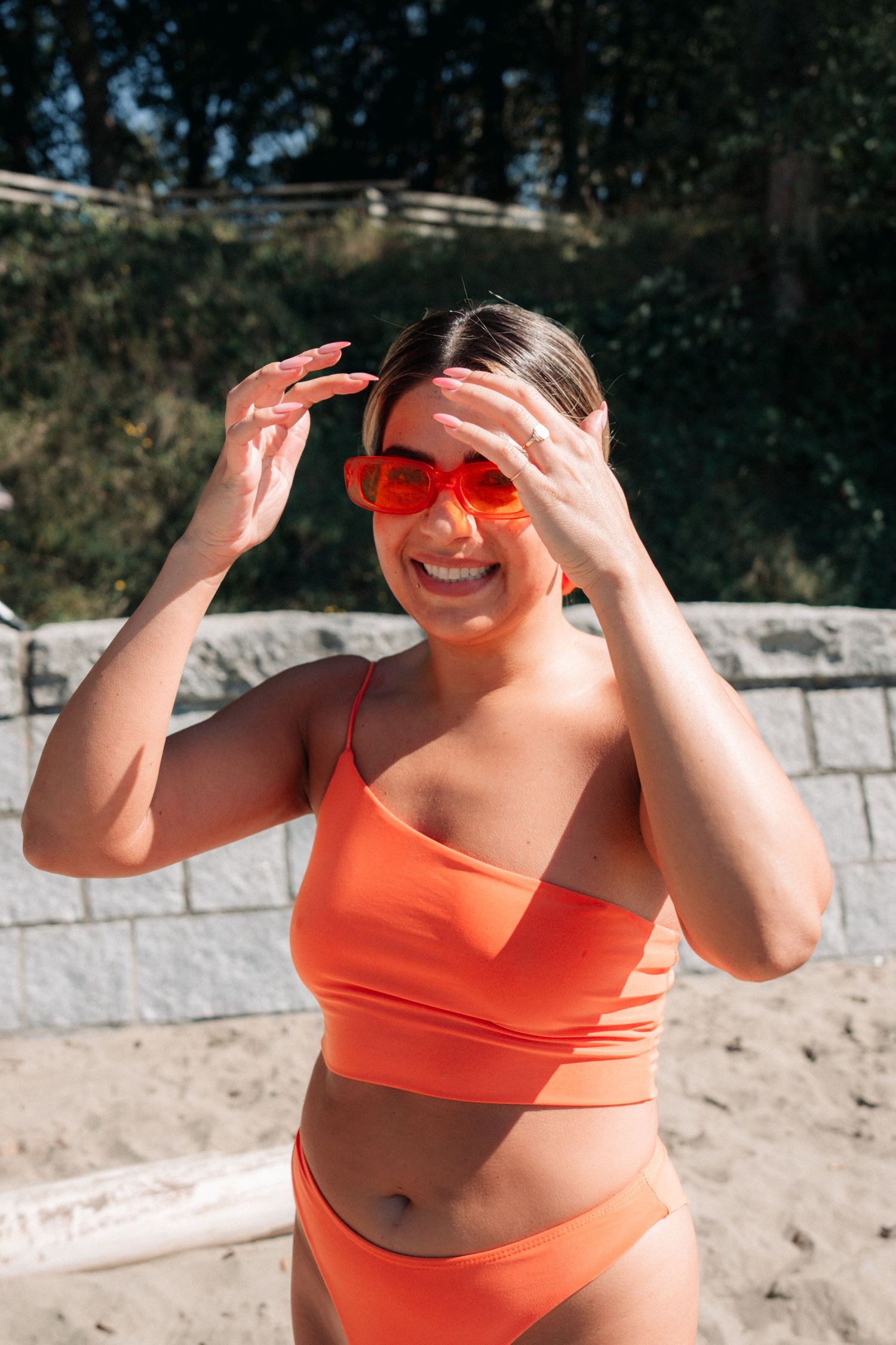Woman in bright orange swimwear and sunglasses shielding face from sun on beach.