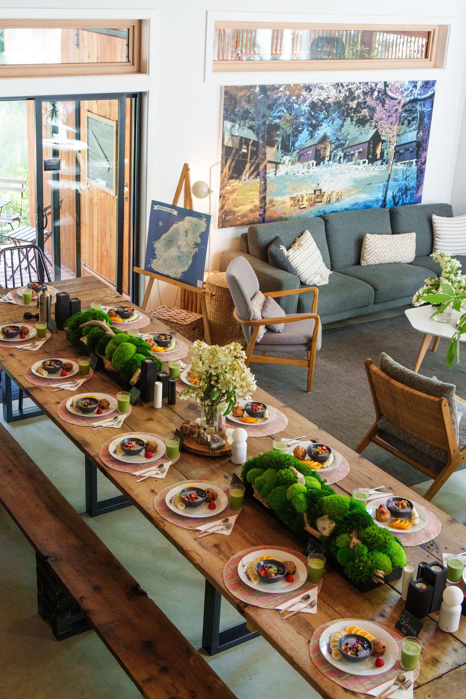 Elegant dinner table setting with green moss decor centerpieces and colorful wall art in a modern dining space.