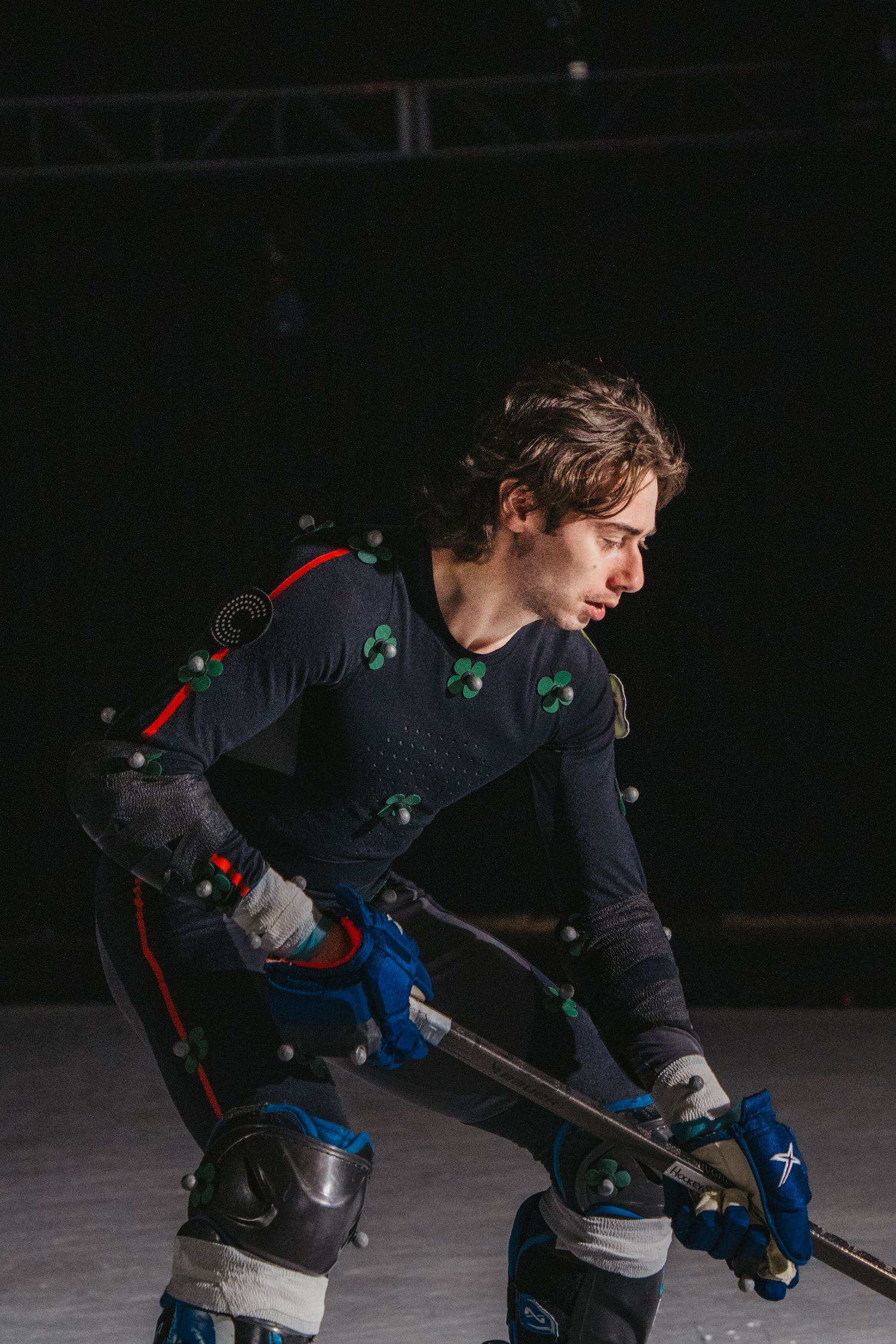 Athletic figure in motion capture suit with tracking markers performs action sequence in dark studio setting.