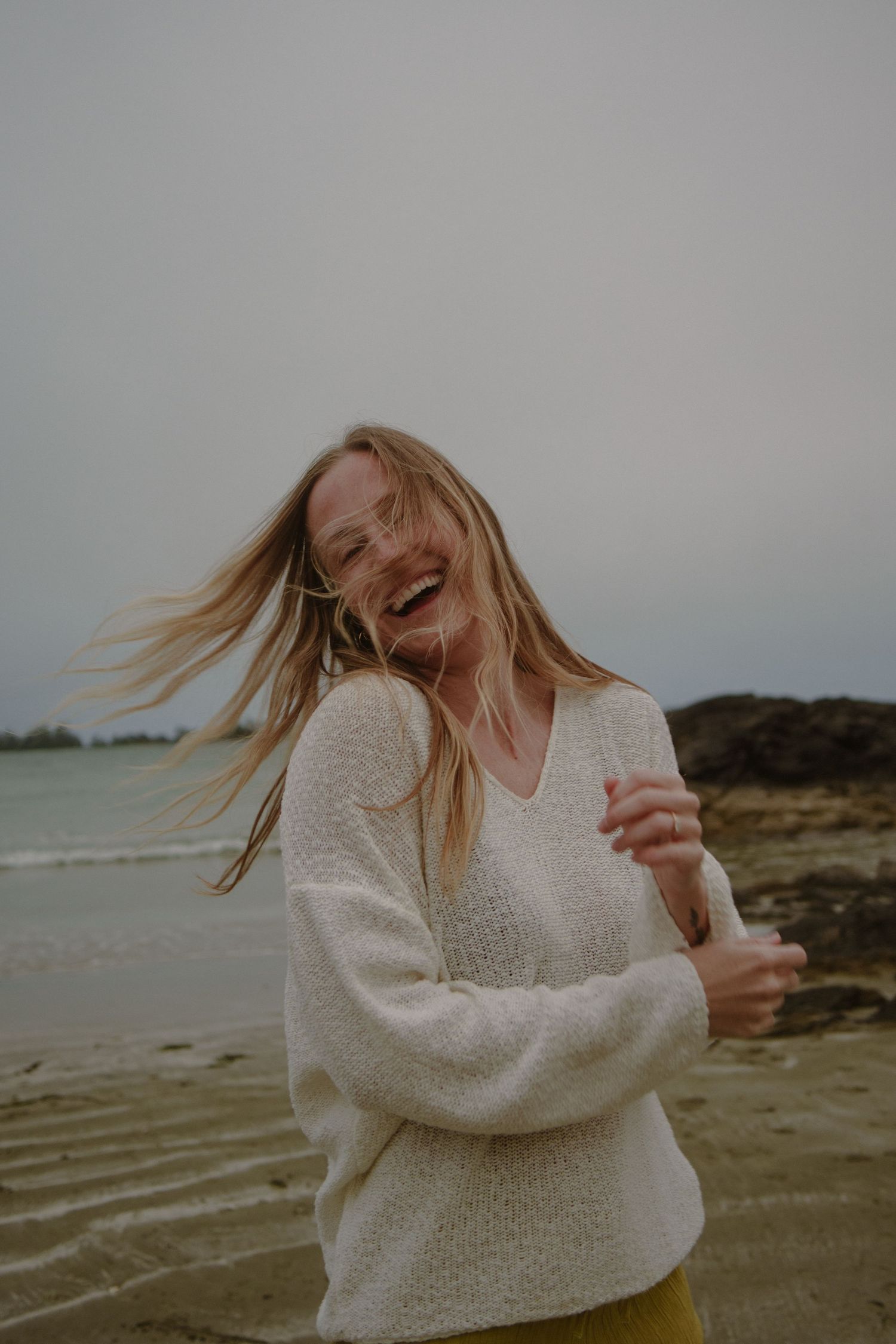 Woman in white sweater laughing joyfully at beach during golden hour.