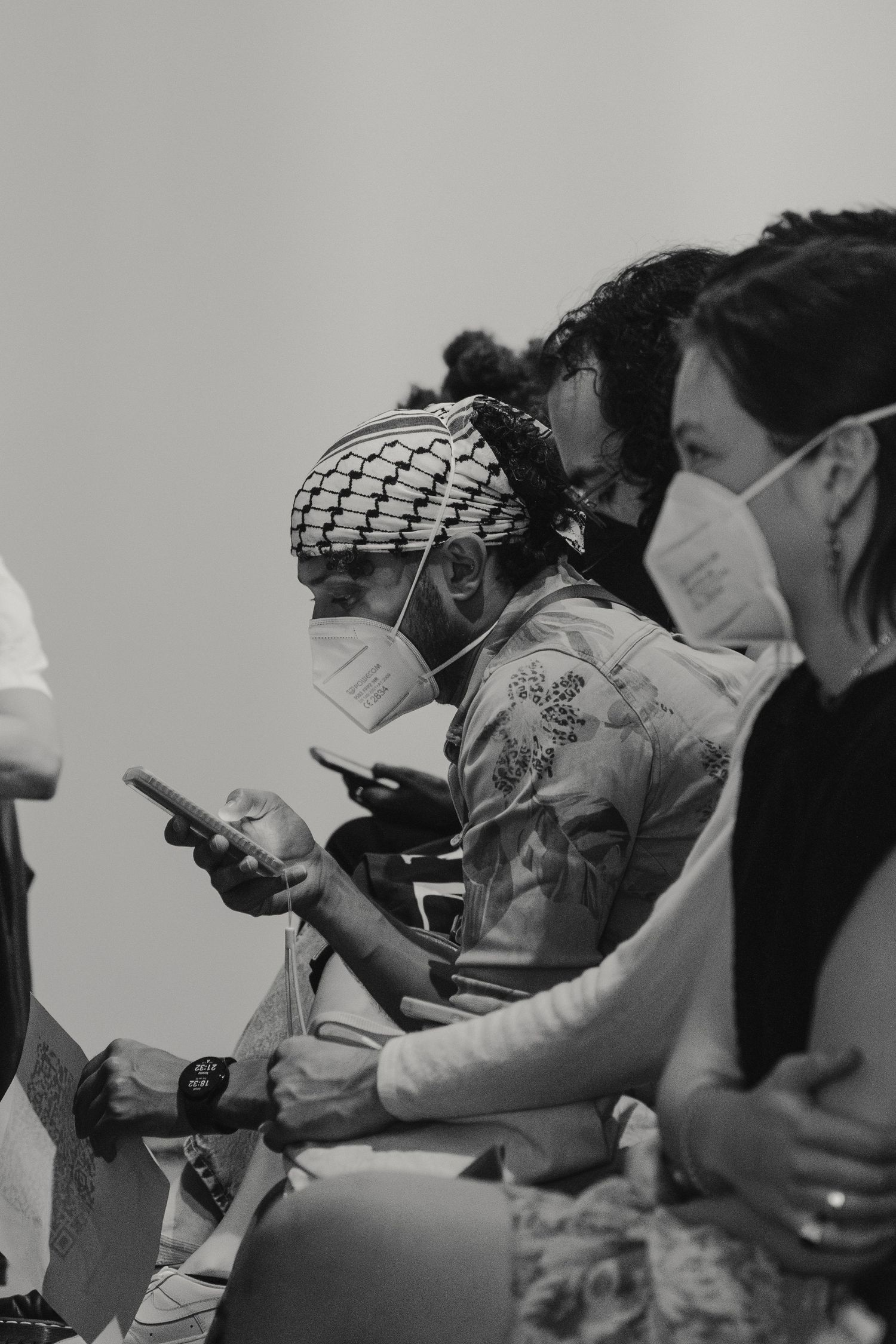 Audience members in masks check phones during indoor gathering in black and white photo.