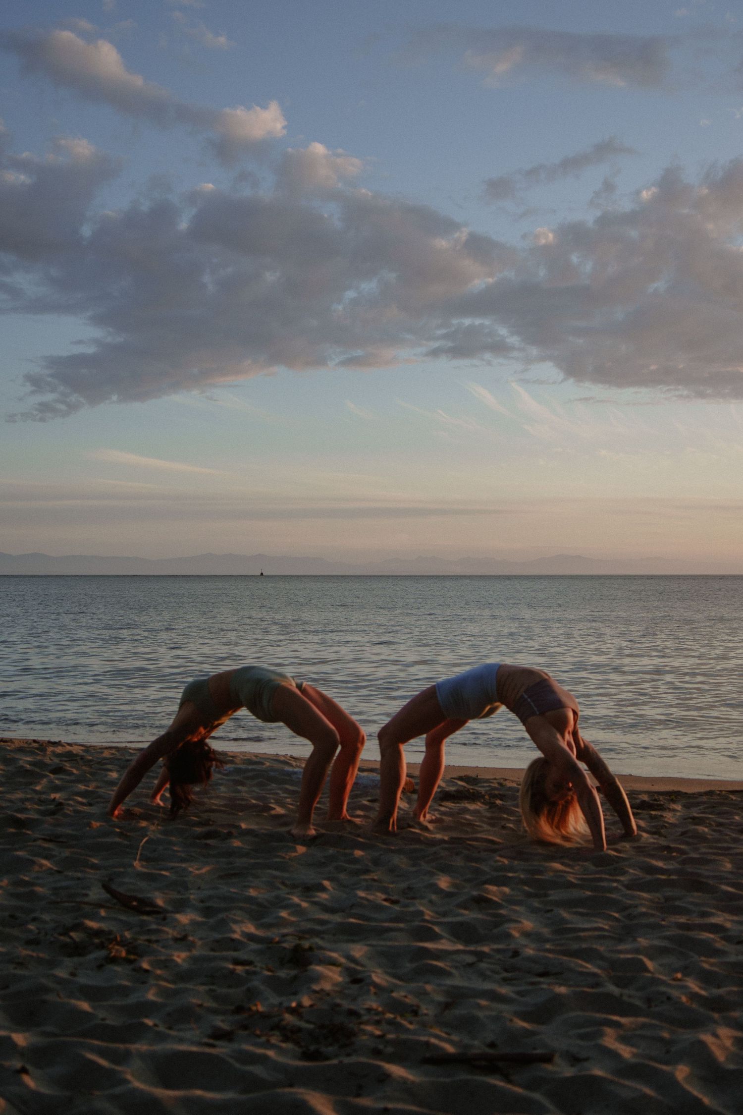 Two people performing synchronized backbend poses on beach at dusk.