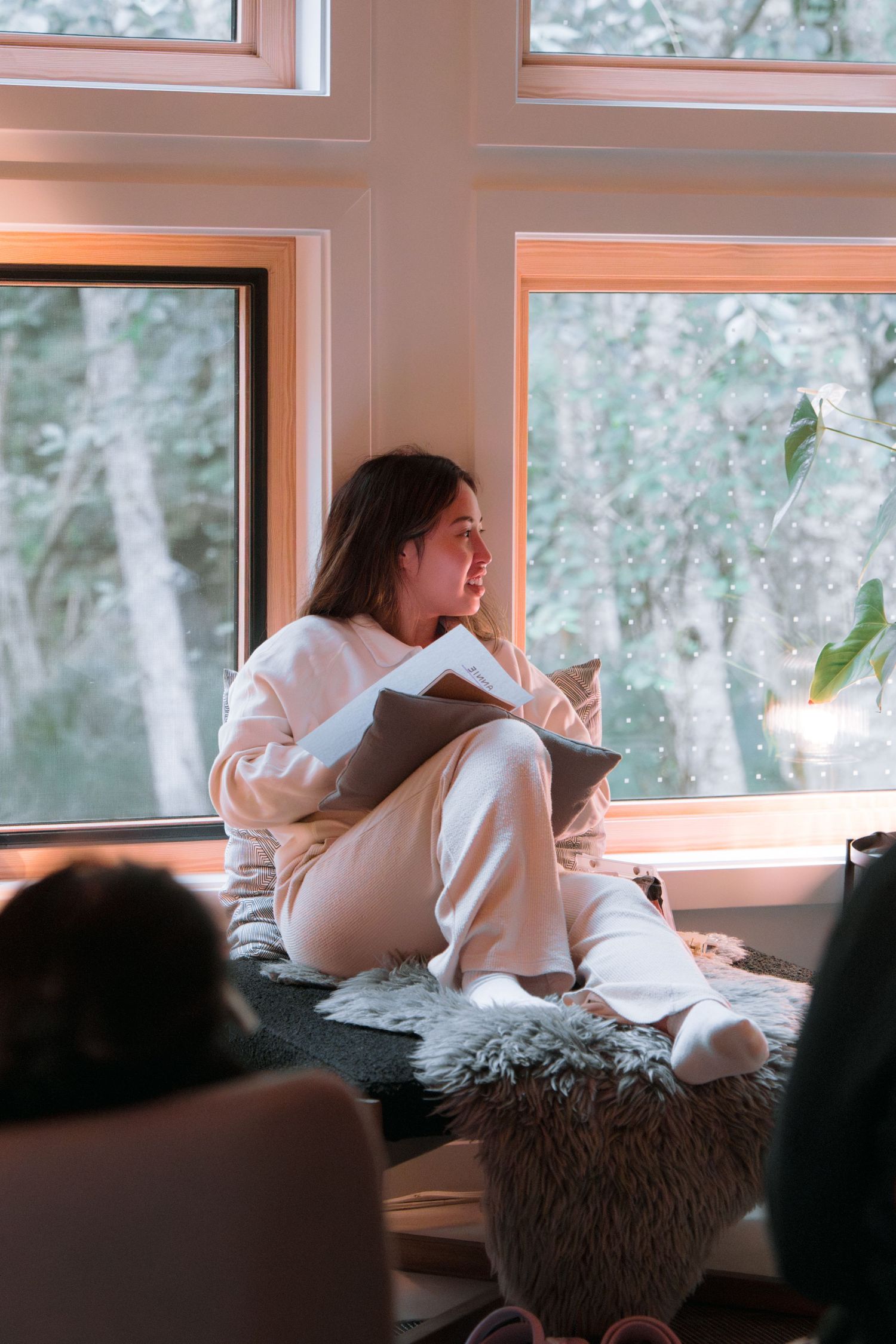 Someone sitting by large windows with a notebook in a cozy reading nook.