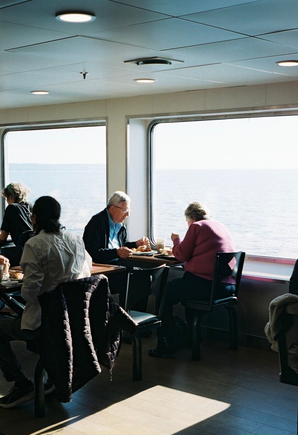 Passengers dining at tables on ferry with ocean view through large windows.