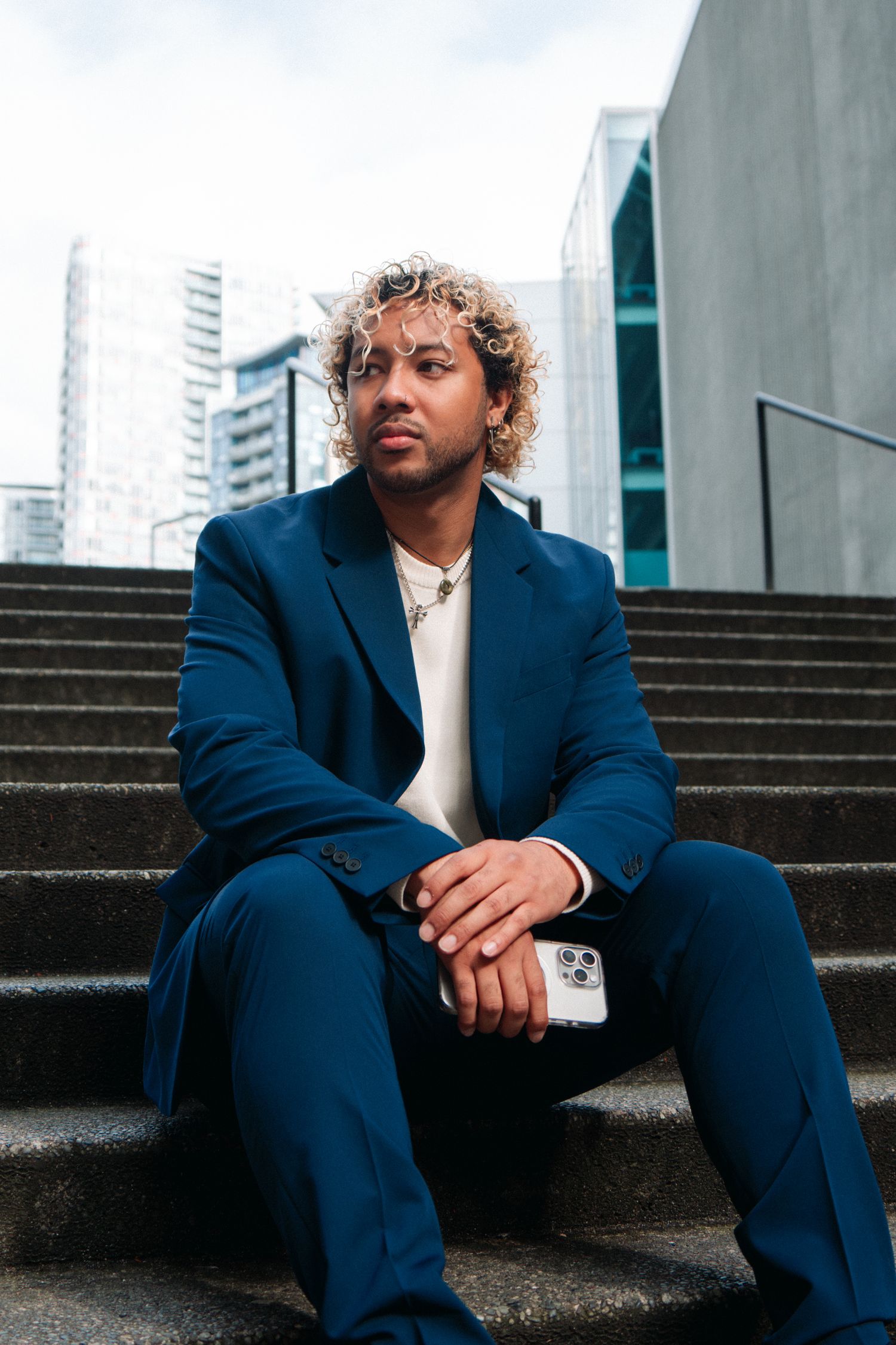 Professional portrait of someone in a navy blue suit sitting on concrete stairs with urban architecture in background.
