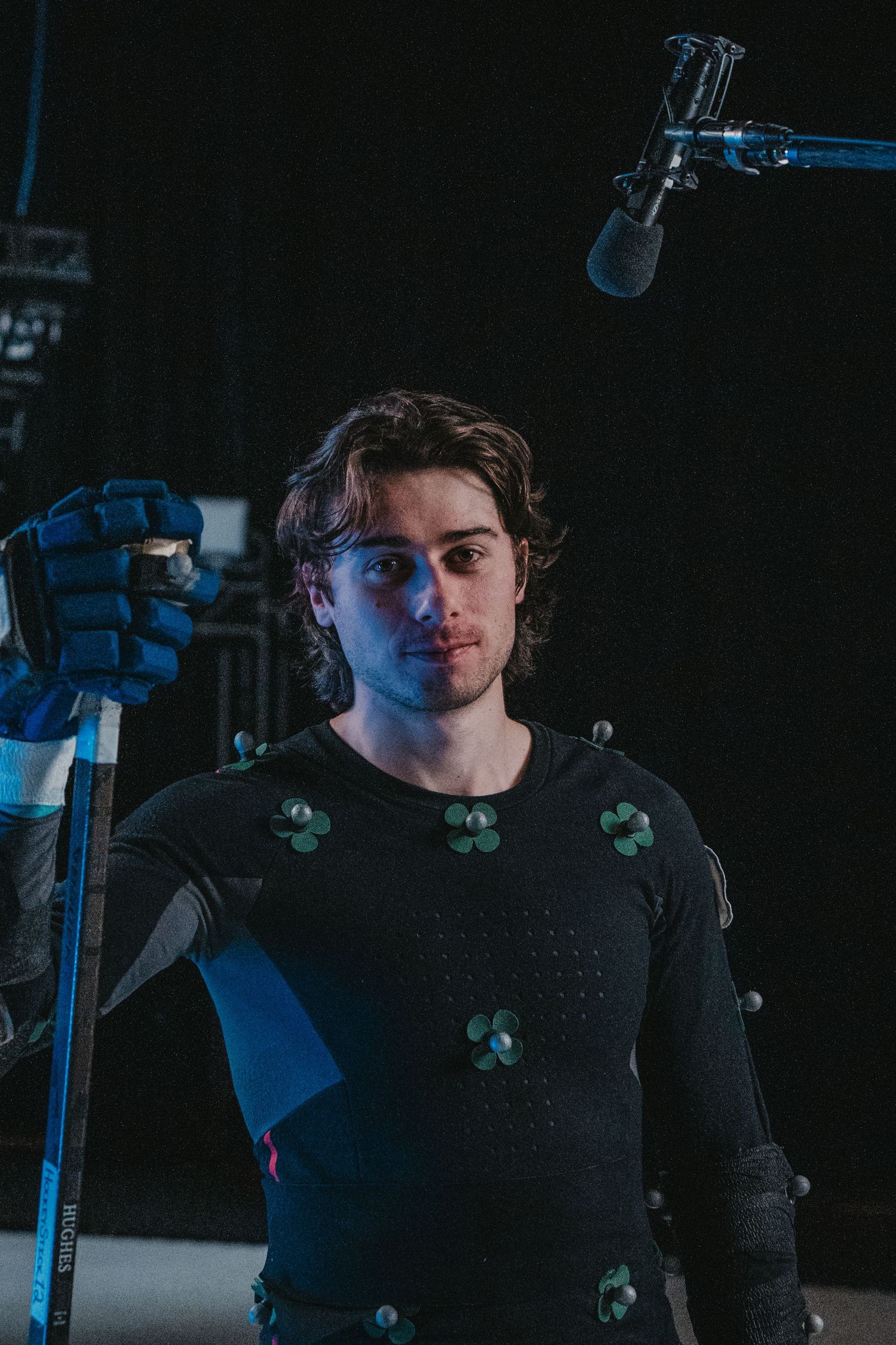 Motion capture actor with blue gloves poses near microphone in dark studio setting.
