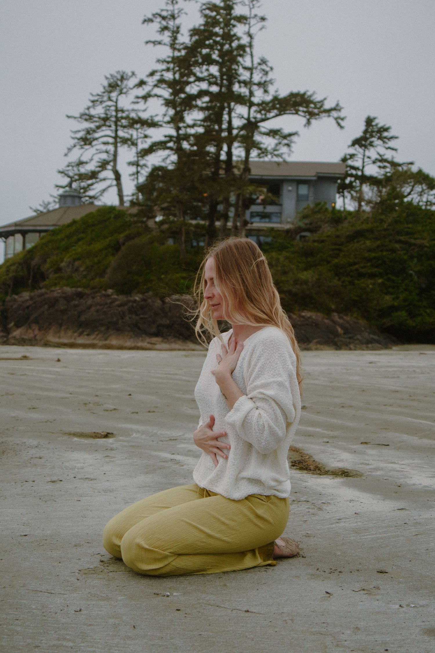 Person practicing meditation on a misty beach with tall evergreen trees and a house on a cliff in the background.