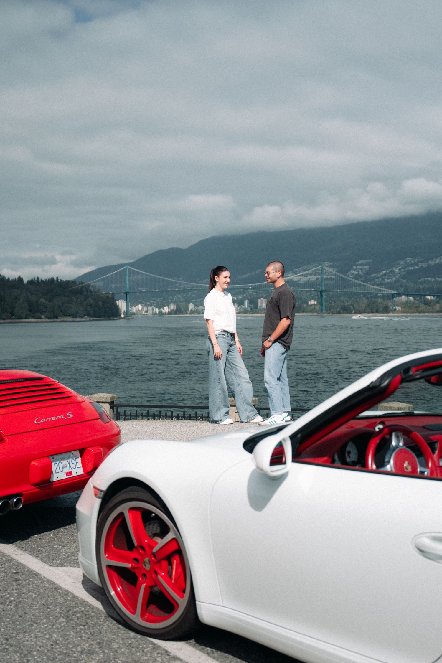 Two sports cars parked at a scenic waterfront location with mountains and bridge in the background.